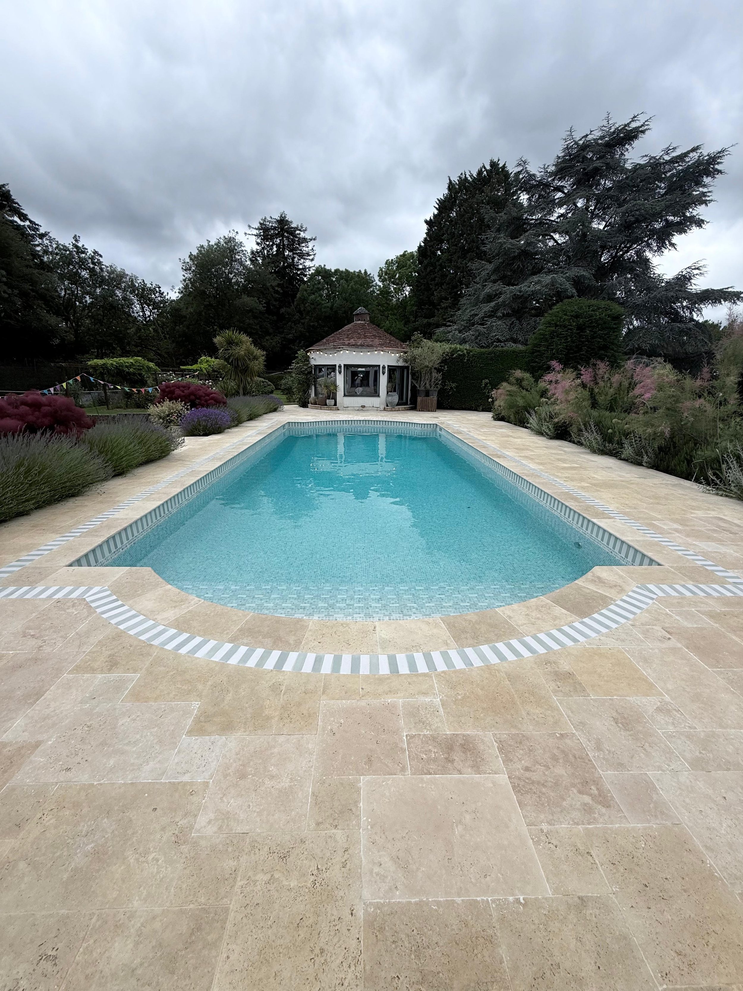 Swimming pool with beige stone tiles surrounding it, a small white building with a conical roof in the background, and lush greenery and tall trees under a cloudy sky.