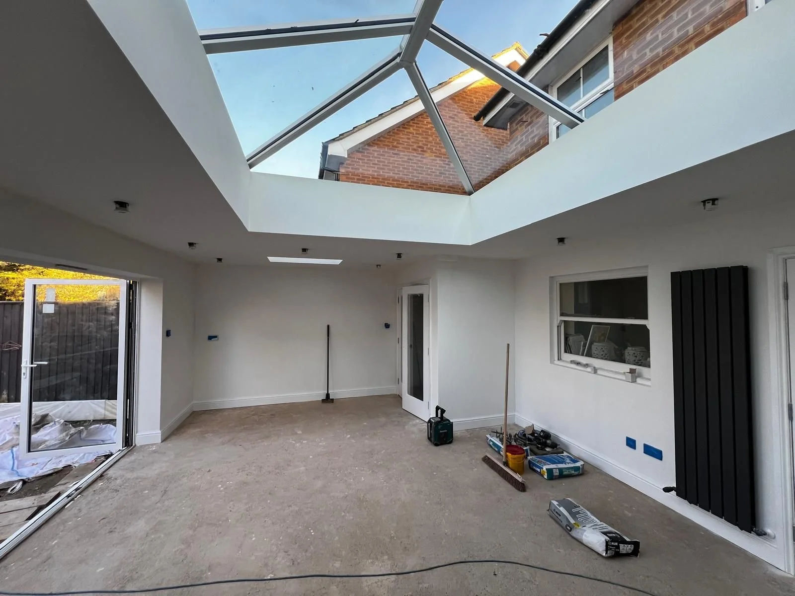Interior of a house under renovation, with a large glass ceiling, sliding glass door, and various construction tools and equipment on the floor.