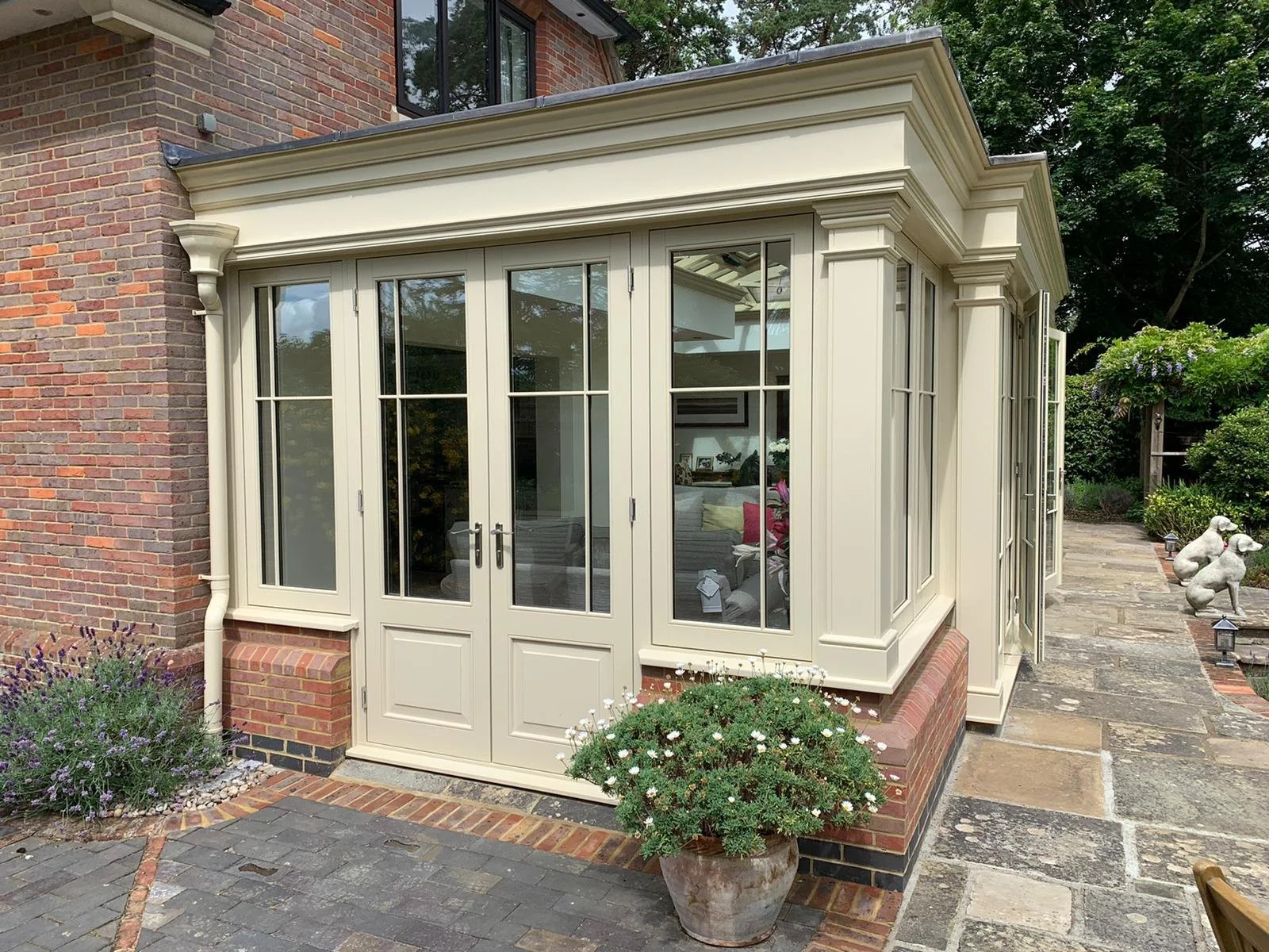 A brick house with a sunroom featuring cream-colored framing, glass doors, and windows, surrounded by potted plants, flowers, and a stone pathway.