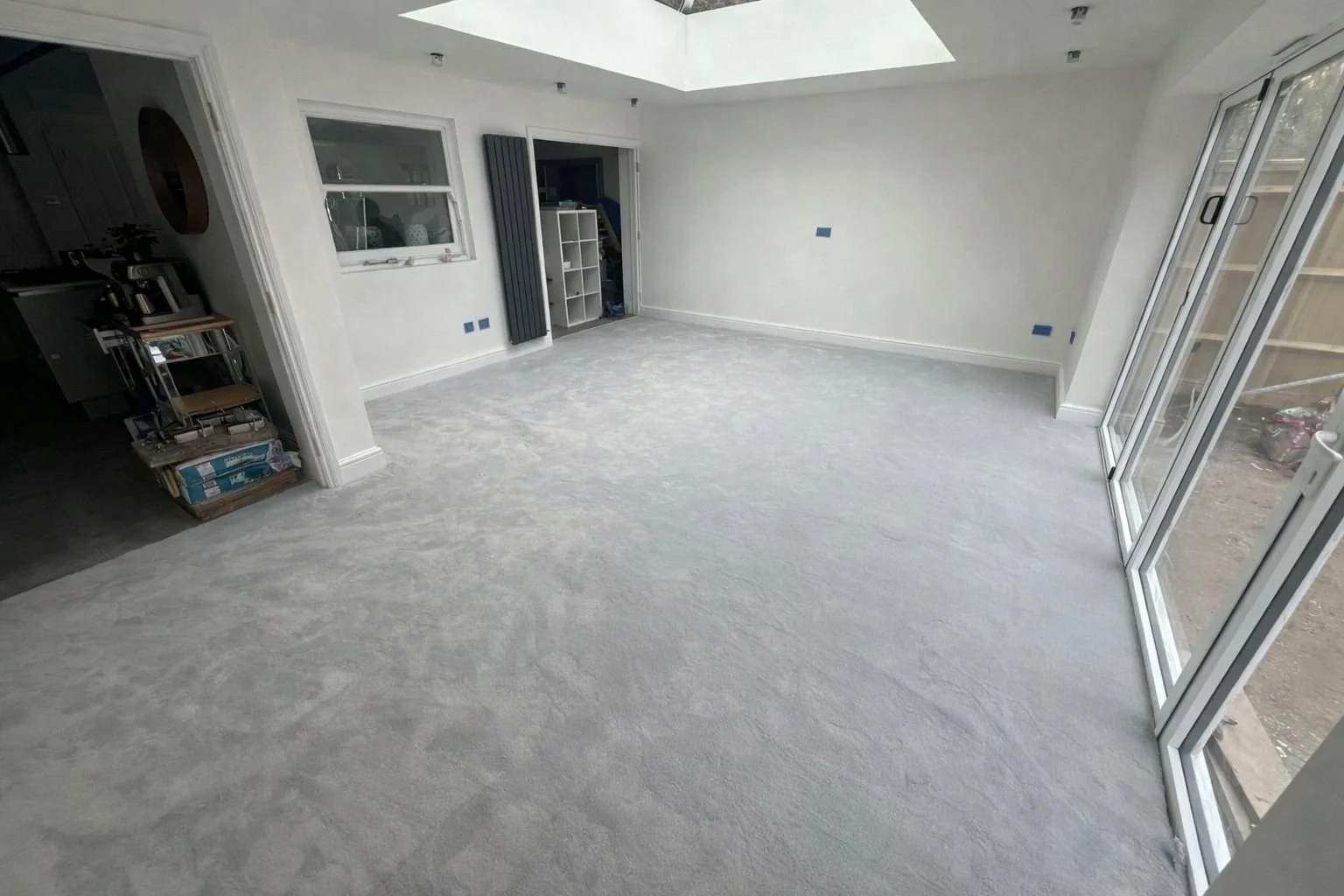 Empty room with large glass sliding doors, white walls, and gray carpet flooring. Some furniture and items are visible in the adjacent room.