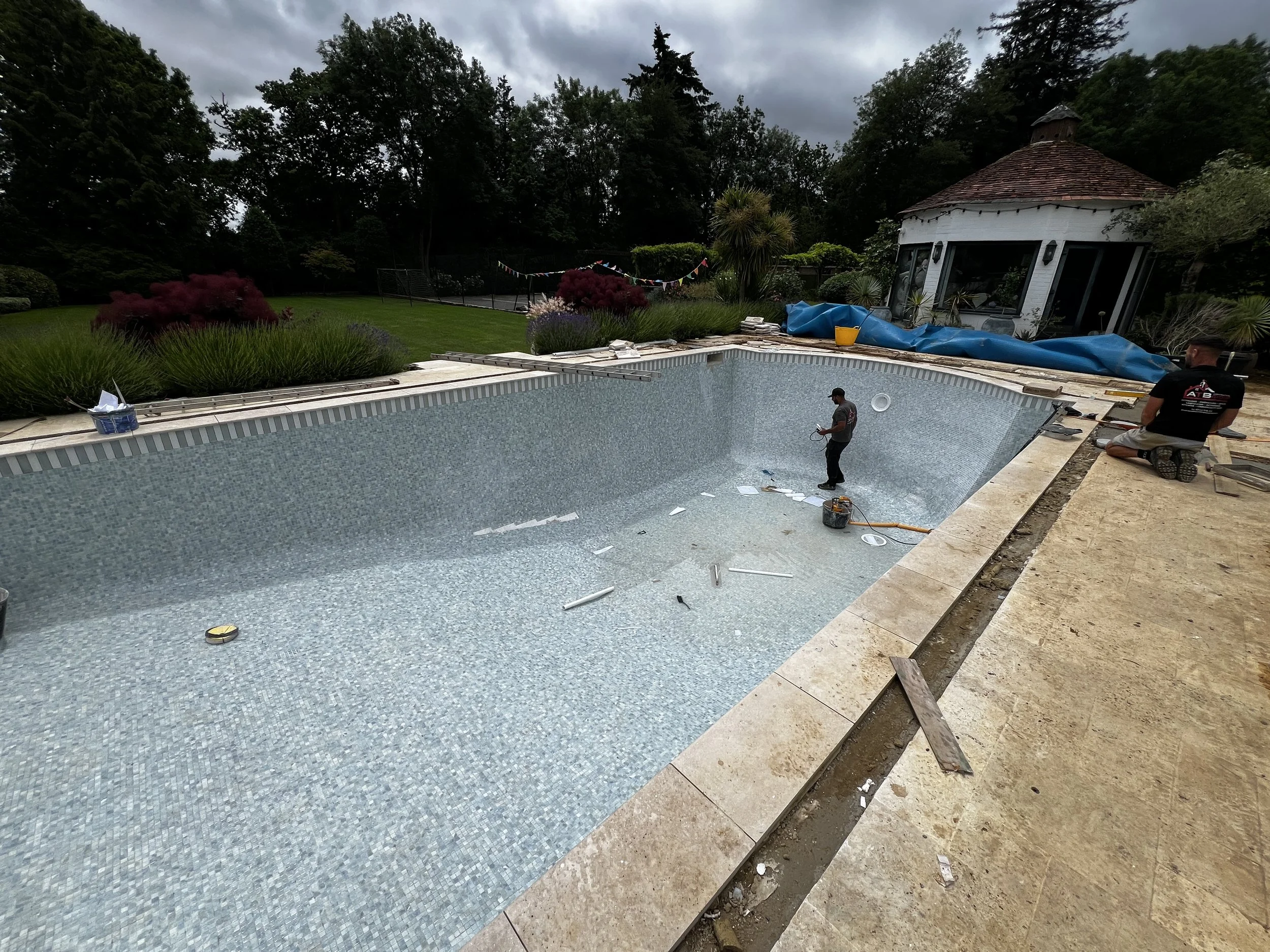 Workers are installing a swimming pool with a blue liner in a backyard, surrounded by landscaping, trees, and a garden house under a cloudy sky.