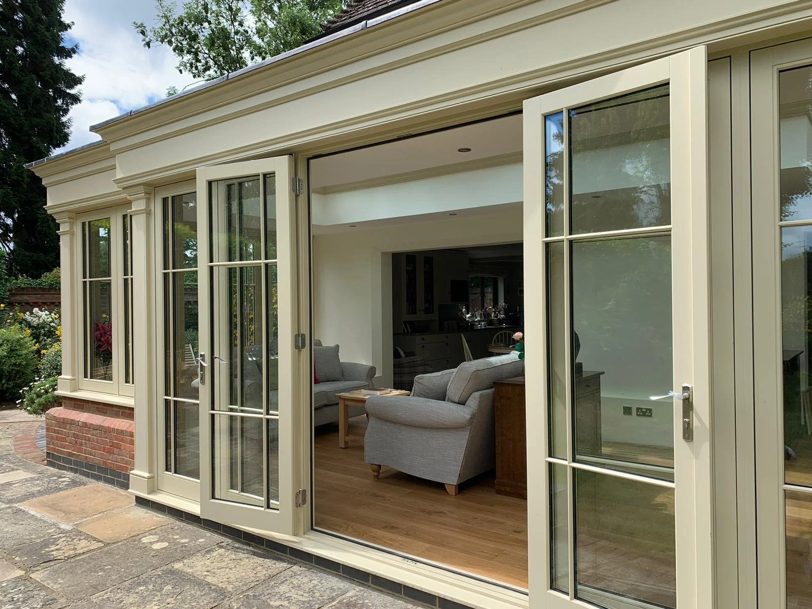 A house with glass doors and windows opening to a patio and a living room with grey sofas, a wooden coffee table, and indoor plants, surrounded by greenery.