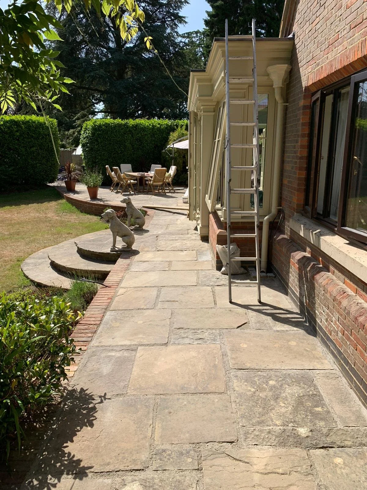 A backyard patio area with stone paving, decorative dog statues, an outdoor dining table with chairs, and a ladder leaning against the house under renovation.