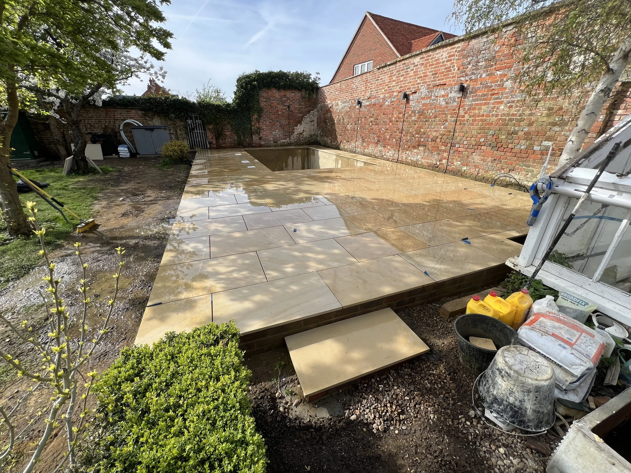 A backyard with a partially constructed patio area featuring large beige tiles. The tiles are wet. There's a brick wall with fencing on top, a small tree, and some garden tools and construction supplies around the area, including bags of mortar and b