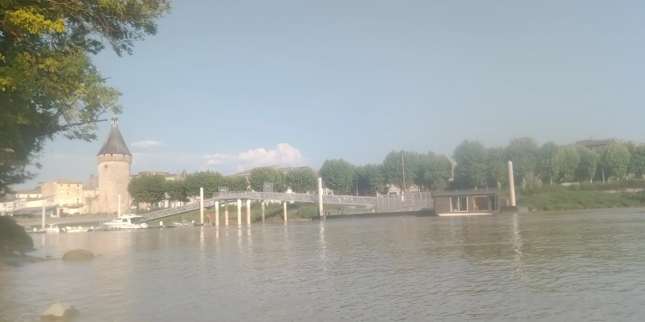 Scène d'une rivière avec le sauna boat libournais amarré au pont piétonnier de Libourne, des bâtiments et un château avec une tour, entourés d'arbres, sous un ciel bleu avec quelques nuages.