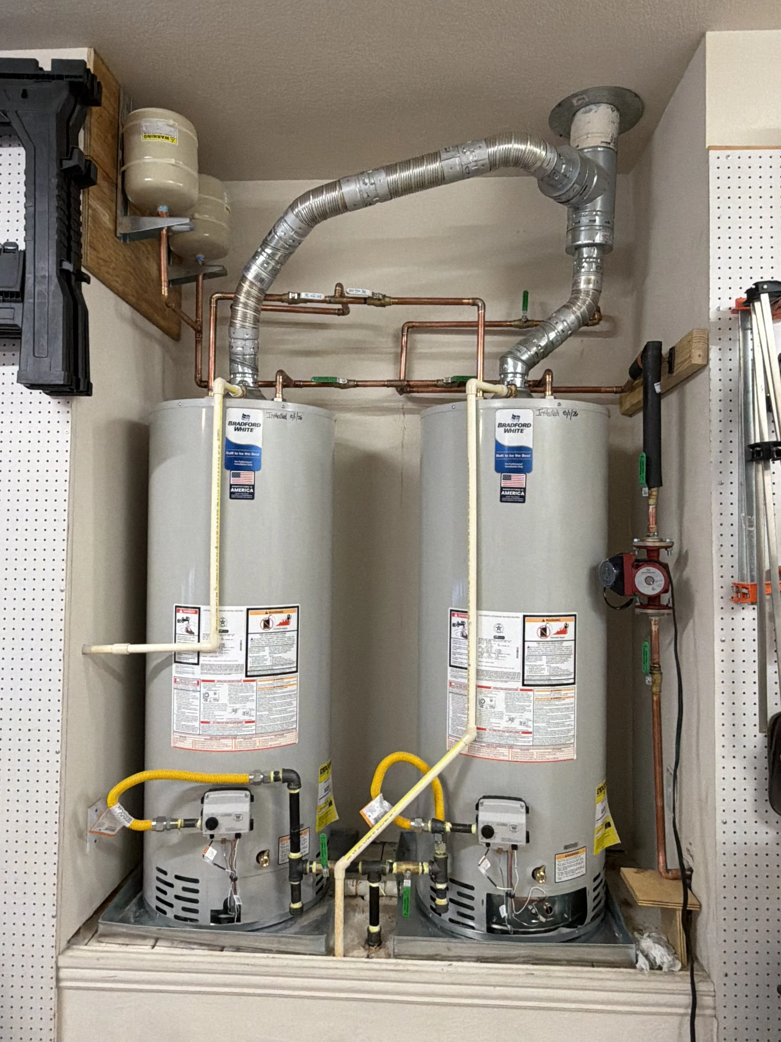 Two water heaters installed side by side in a utility closet, connected by pipes and valves, with a vent pipe on top.