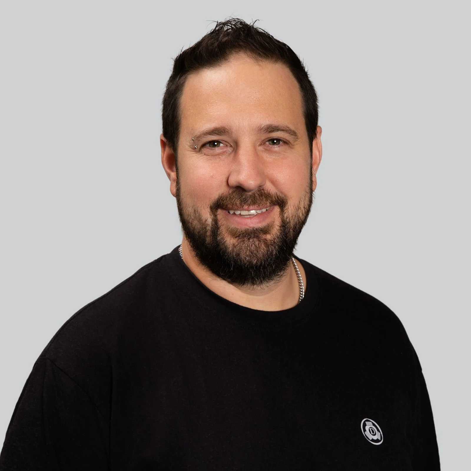 Ein Mann mit dunklem Haar, Bart und Piercing im rechten Augenbrauen, trägt ein schwarzes T-Shirt mit einem Logo auf der linken Seite.