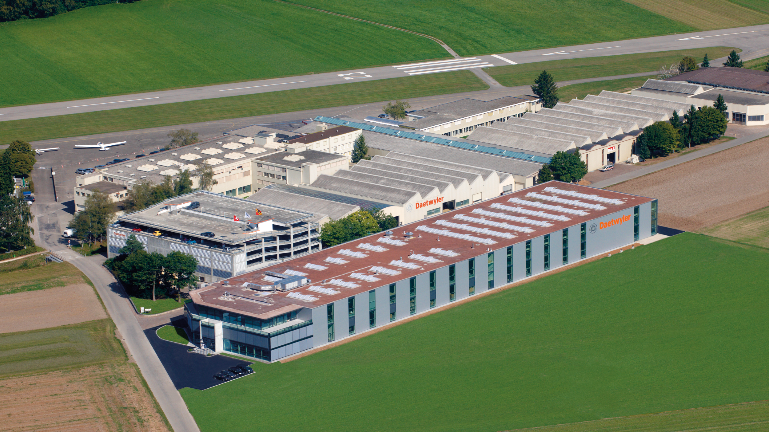 Luftaufnahme eines Gewerbegebäudes mit Firmenlogo Daetwyler, umgeben von Feldern, ein Parkplatz mit Autos und Flugzeuge neben dem Gebäude.