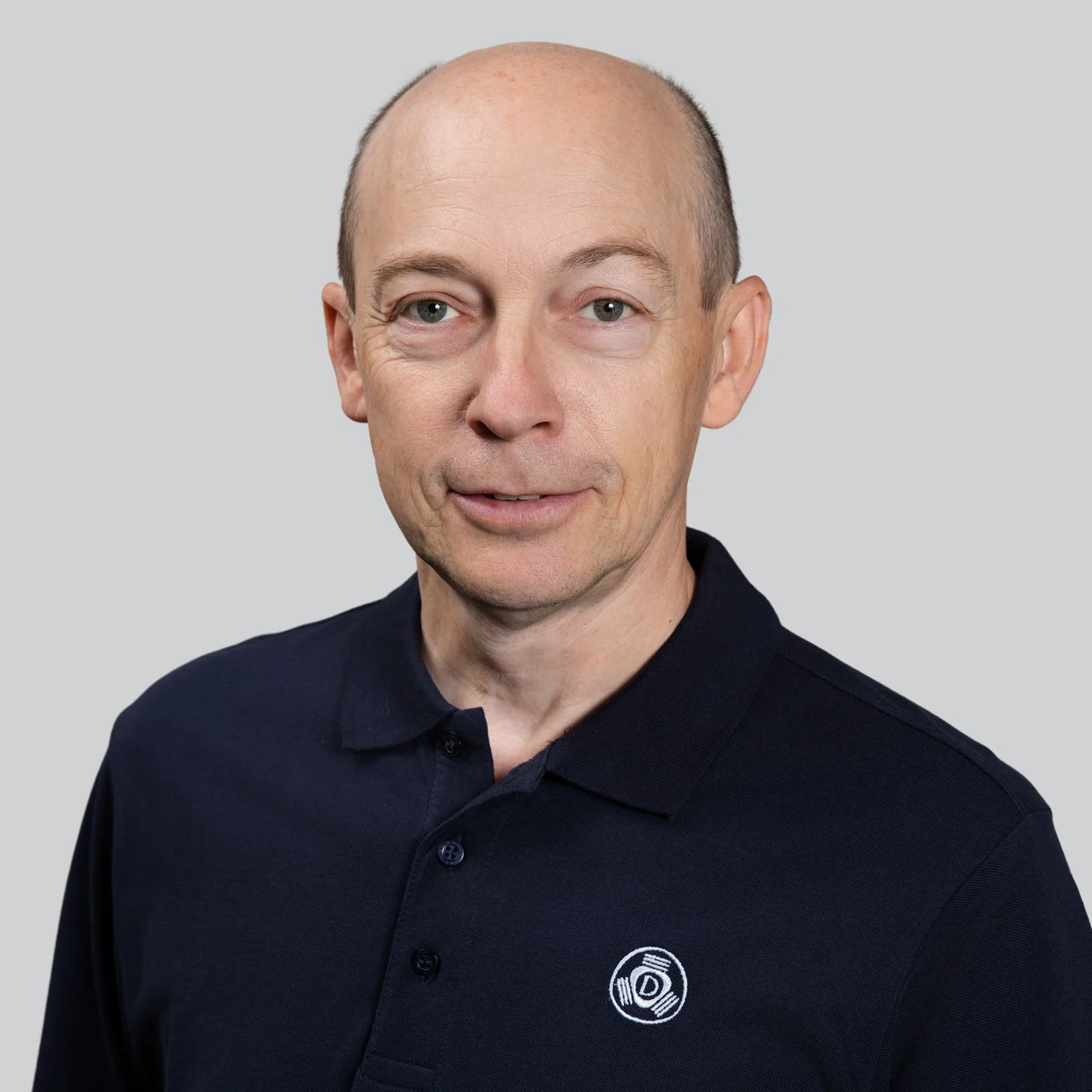 Ein Mann trägt ein schwarzes Polo-Shirt mit einem weißen Logo auf der linken Brustseite, glattem Kopf, blauen Augen, vor einem hellgrauen Hintergrund.
