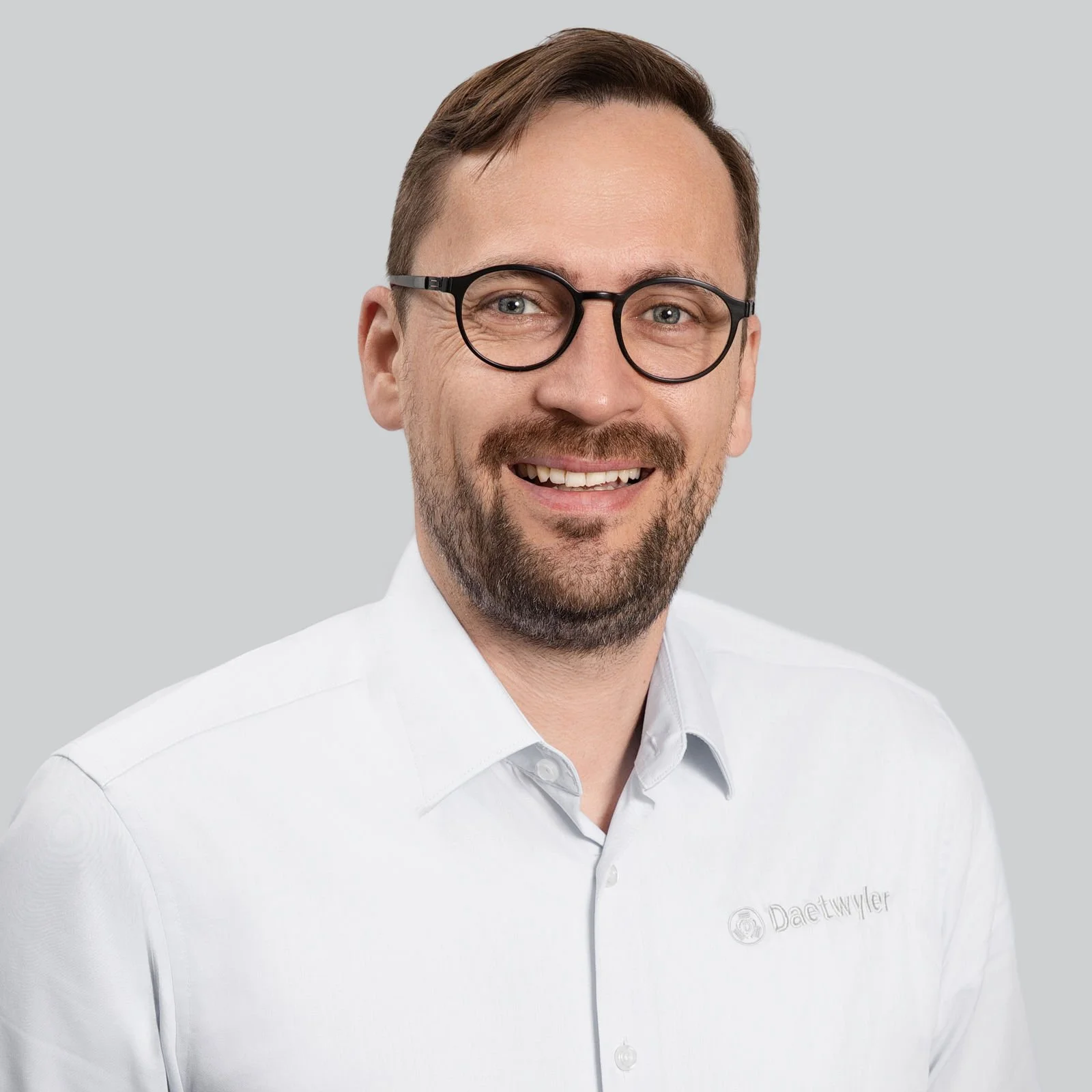 Porträt eines Mannes mit Brille, kurzer brauner Haar- und Bartfrisur, lächelnd, trägt einen weißen Hemd mit Logo "Daetwyler" auf der Brust, grauer Hintergrund.