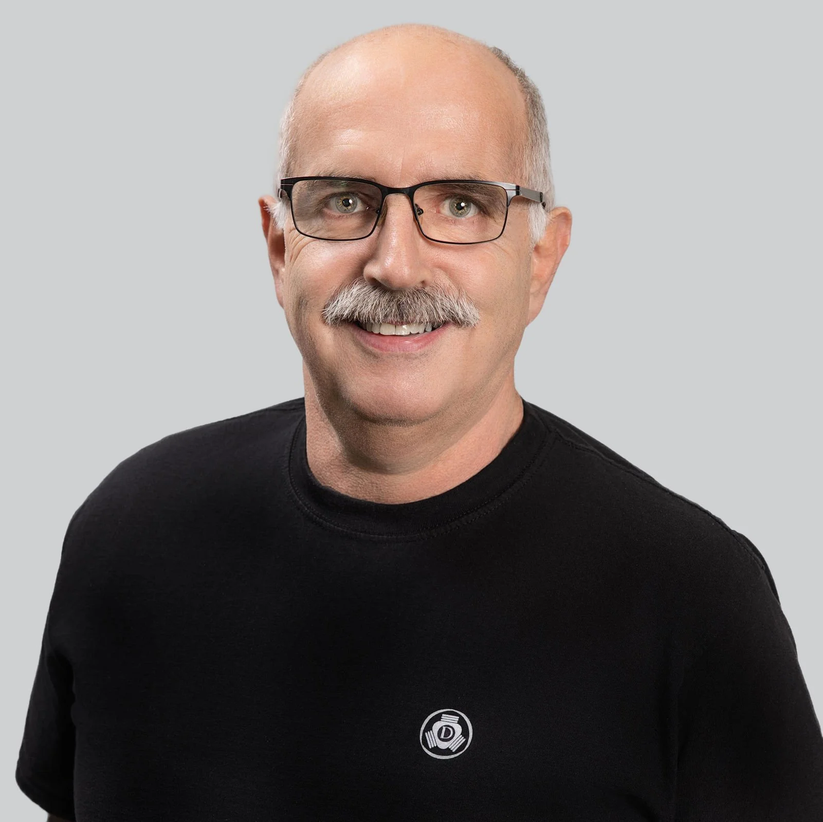 Ein Mann mit Brille, grauem Bart und aufgeschossenem Kopf trägt ein schwarzes T-Shirt mit einem Logo, das eine Hantel und den Buchstaben 'D' zeigt.