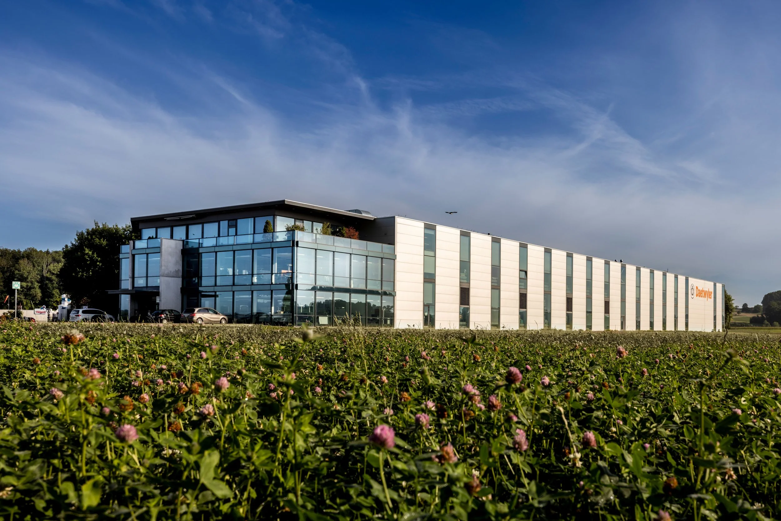 Moderne Bürogebäude mit Glasfassade, umgeben von Grünflächen und einem blauen Himmel.