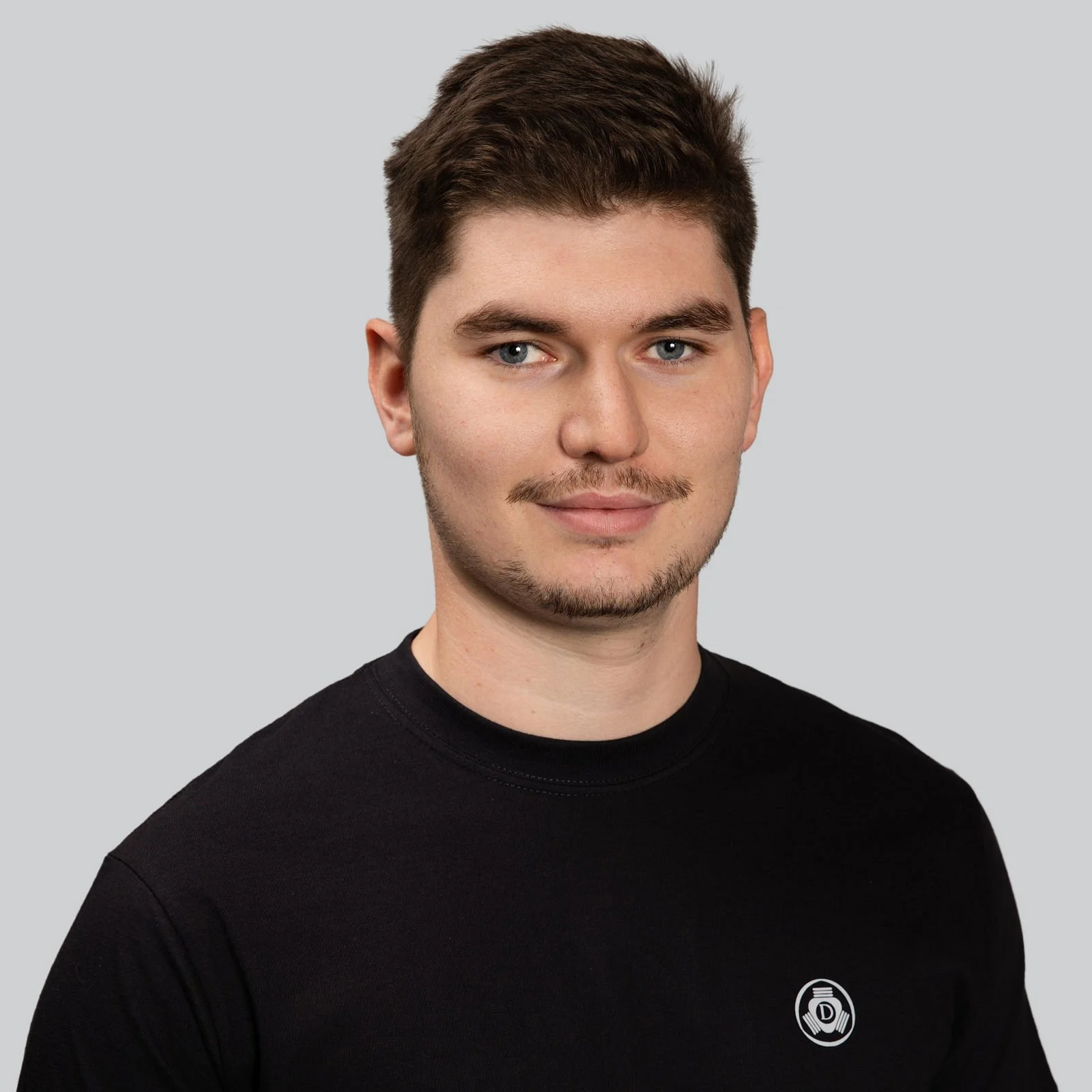 Junger Mann trägt schwarzes T-Shirt mit einem Logo auf der linken Brustseite, grauer Hintergrund.