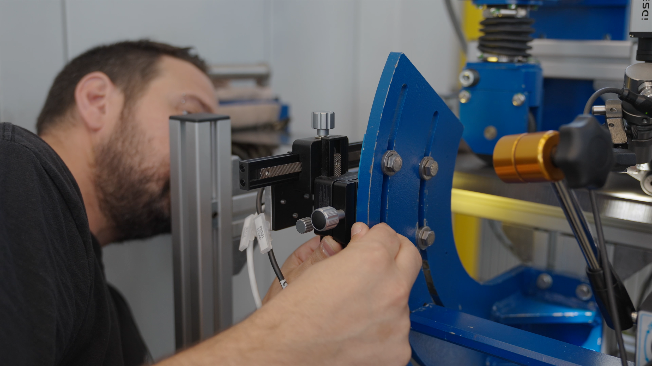Ein Mann arbeitet an einer technischen Vorrichtung in einer Werkstatt oder einem Labor. Er ist konzentriert und benutzt ein Werkzeug zum Justieren oder Fixieren des Equipments, das aus Metallteilen und eine blaue Unterstützung besteht.