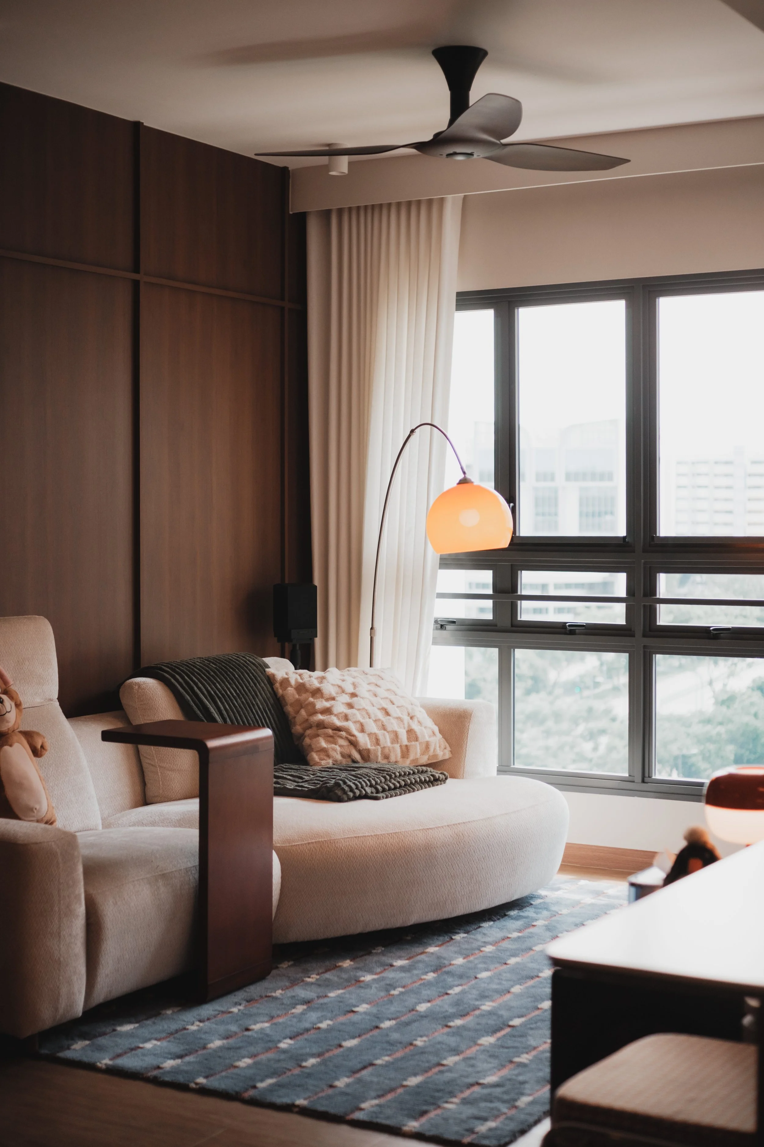 Lucent™ curtains drawn open with sunlight streaming in, in a warm Singapore living room with cosy furniture and ambient lighting.