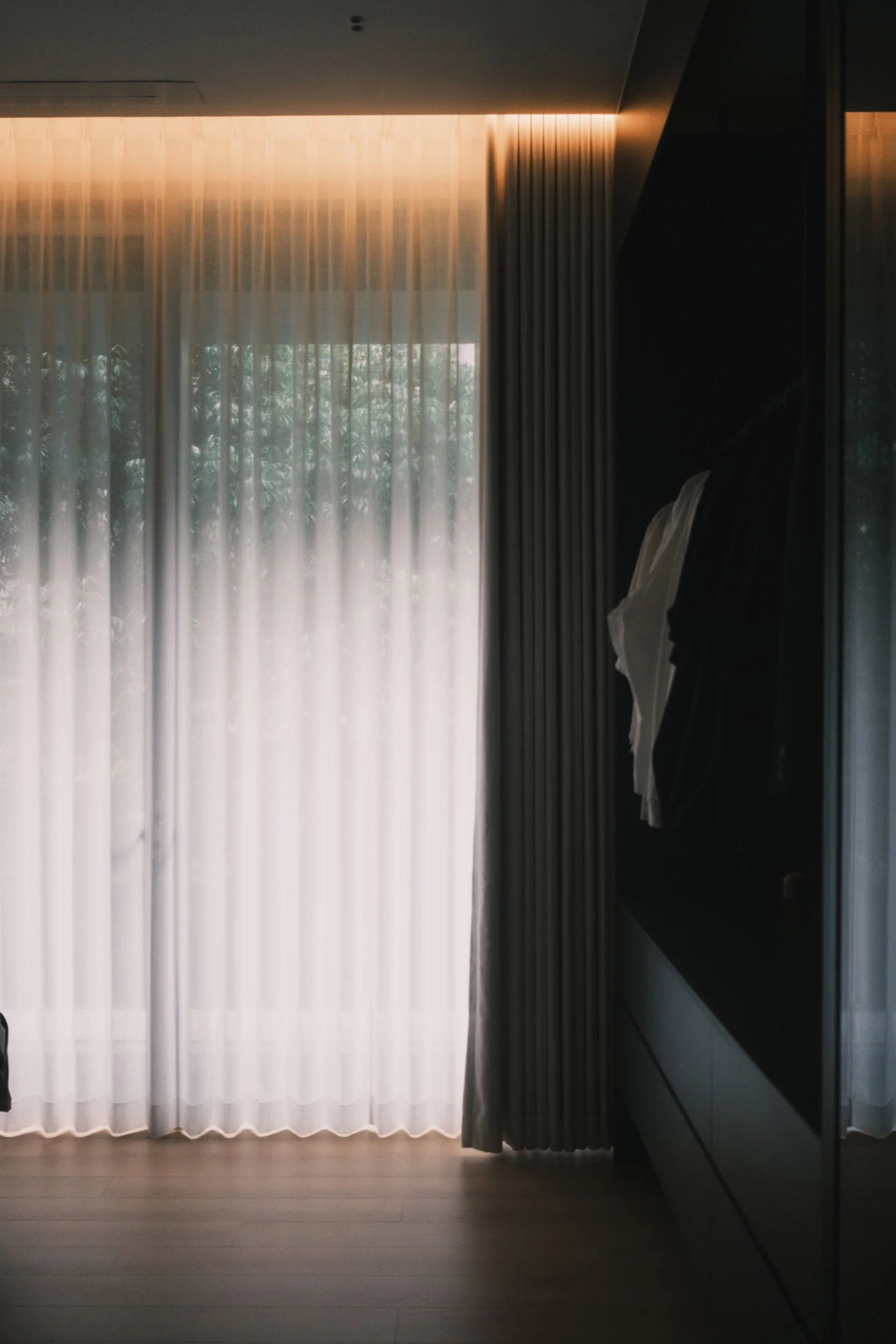 Full-height Lucent and Somn curtains filtering soft natural light beside a dark wardrobe in a moody bedroom interior.