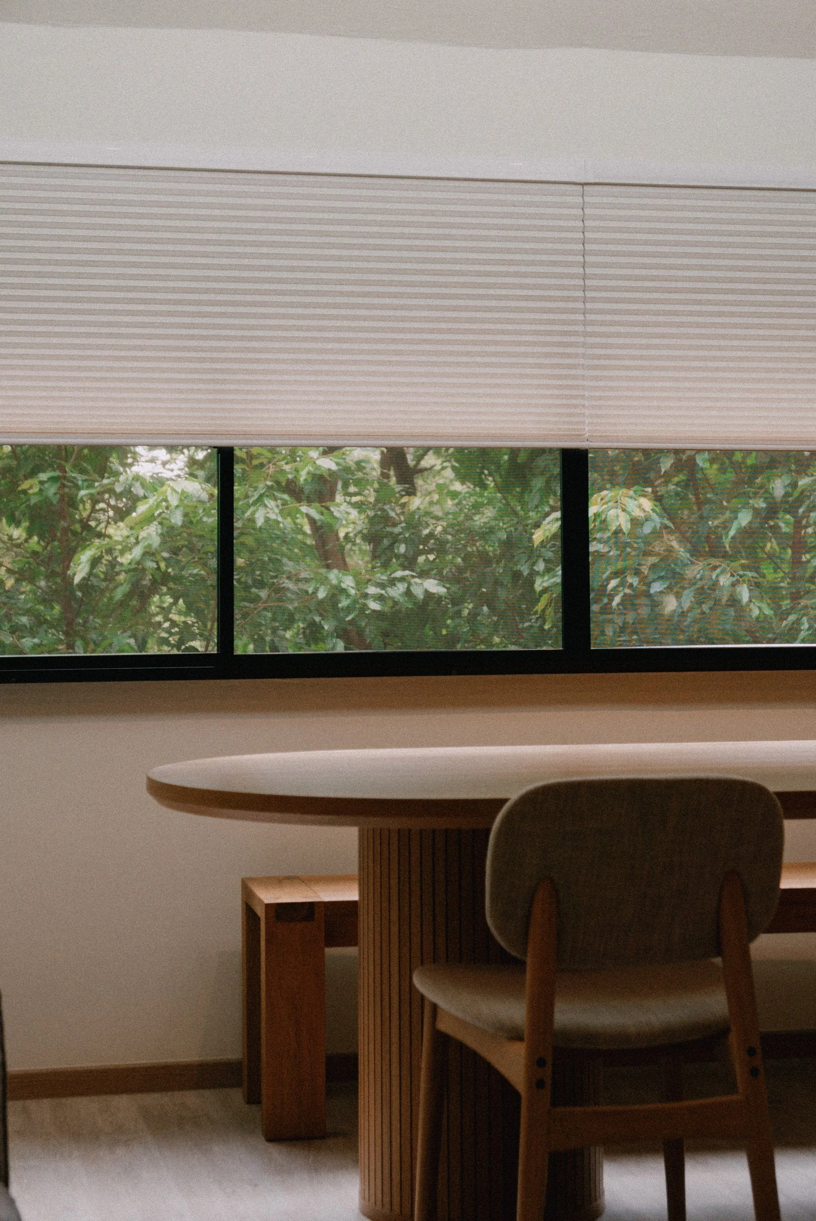 Honeycomb blinds partially covering a wide dining room window, offering privacy while keeping the greenery outside in view.