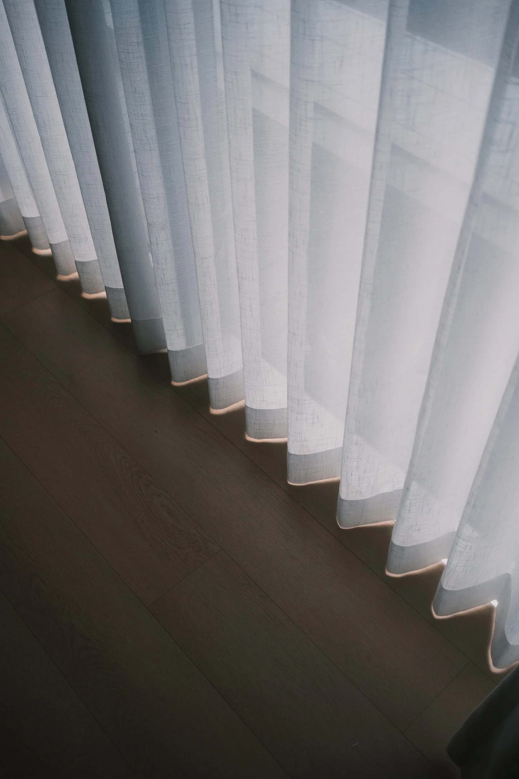 Close-up of Lucent™ sheer curtains softly draping over dark wood flooring, filtering natural light with a gentle, airy finish.