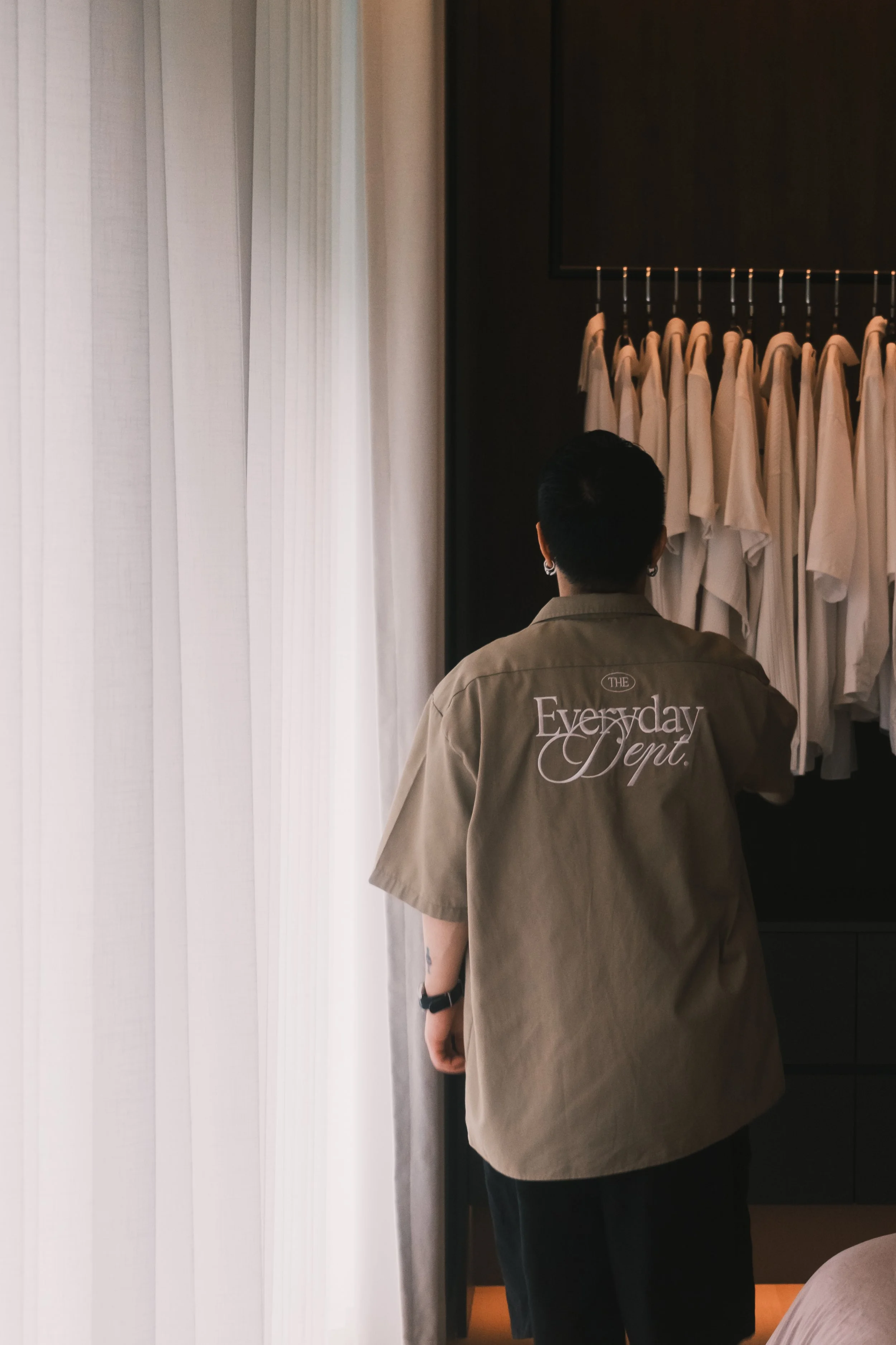 A person with short dark hair, wearing a beige shirt with "Everyday Dept." written on the back, stands facing a closet with a row of white T-shirts hanging on a rack.