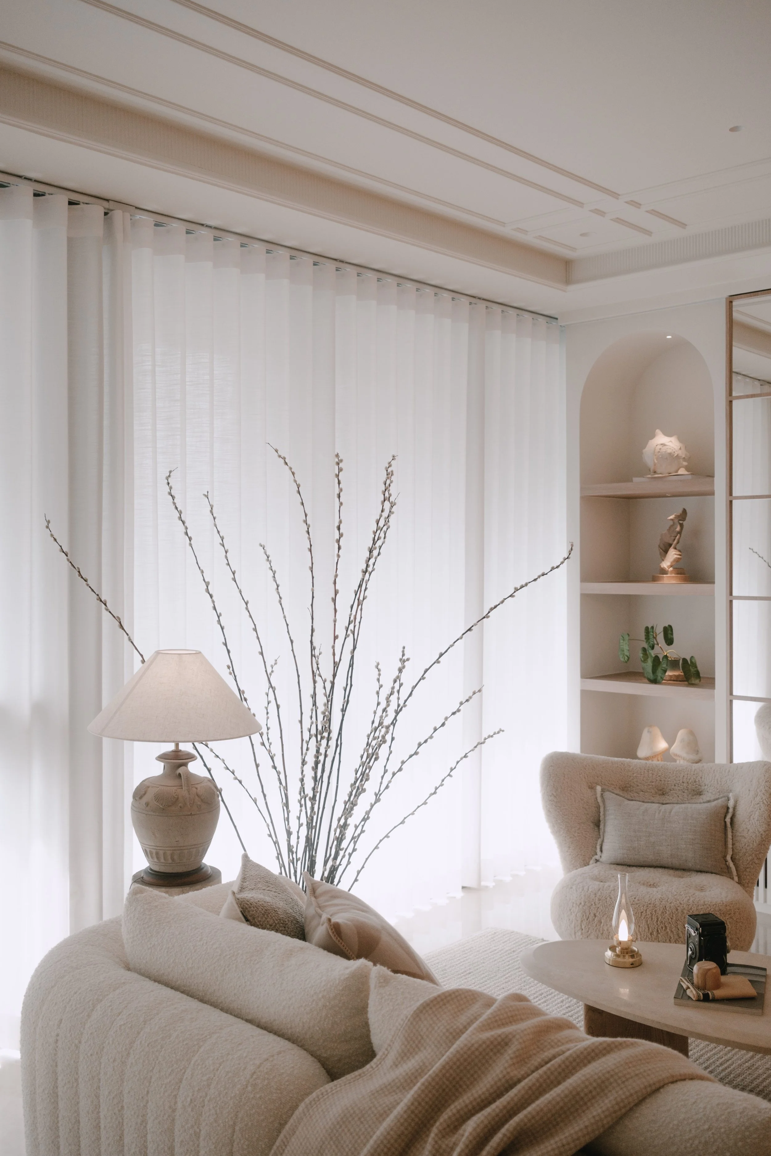 Lucent curtains framing a cosy neutral living corner with soft daylight, an armchair and styled shelving.