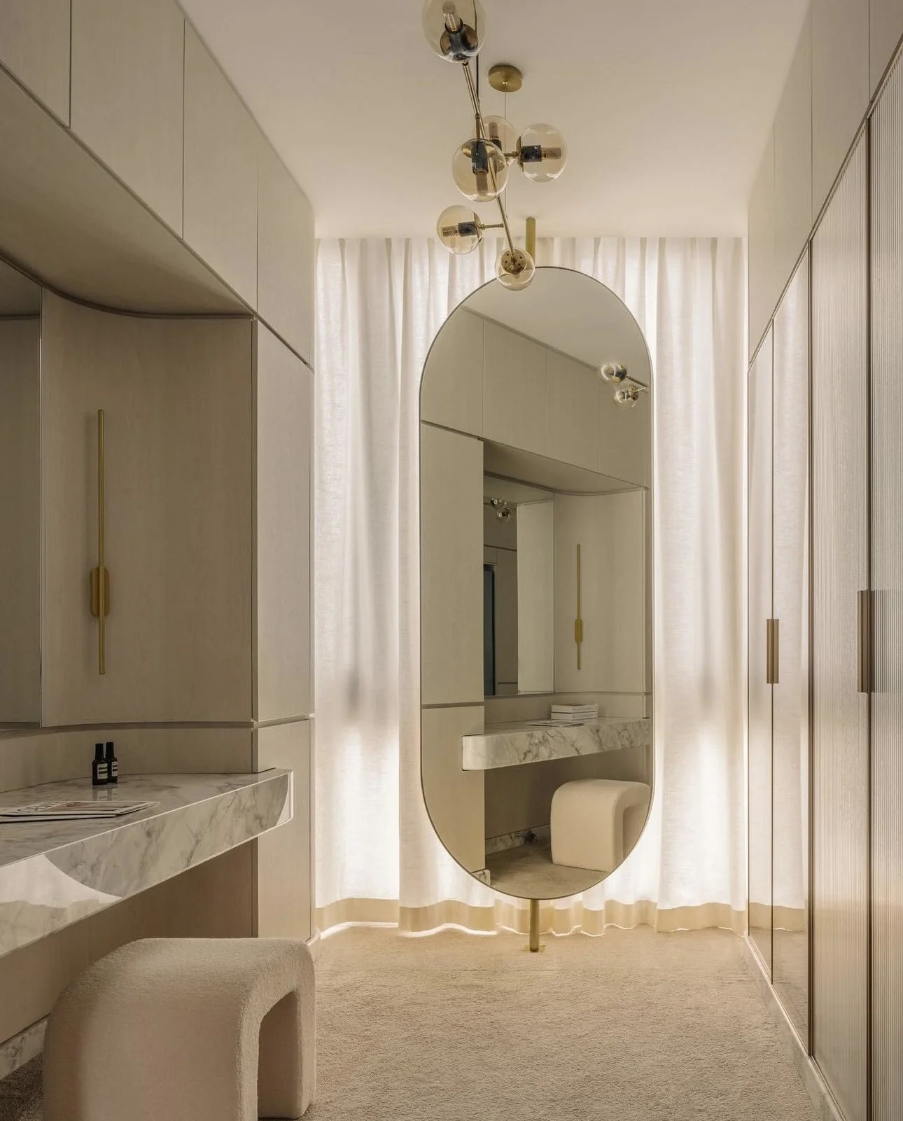 Full-height curtains softly filtering natural light in a walk-in vanity area with a curved mirror, warm neutral tones and built-in wardrobe.