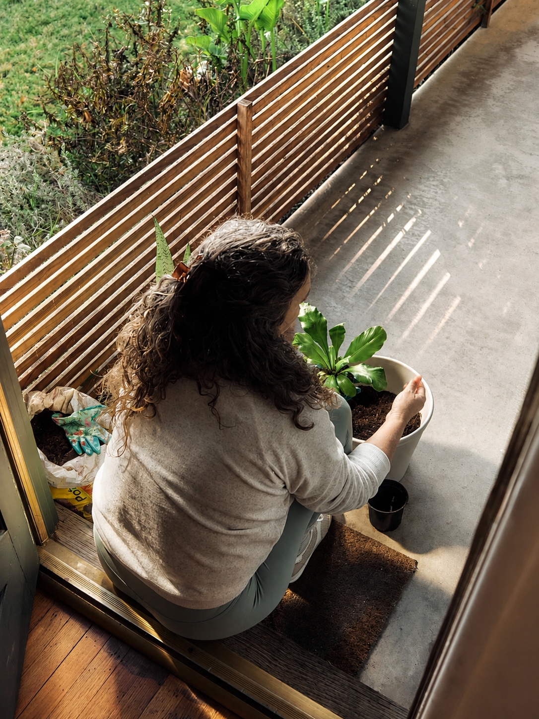 A woman sits on a doorstep in soft daylight, potting a green plant, with a garden and wooden fence behind her.