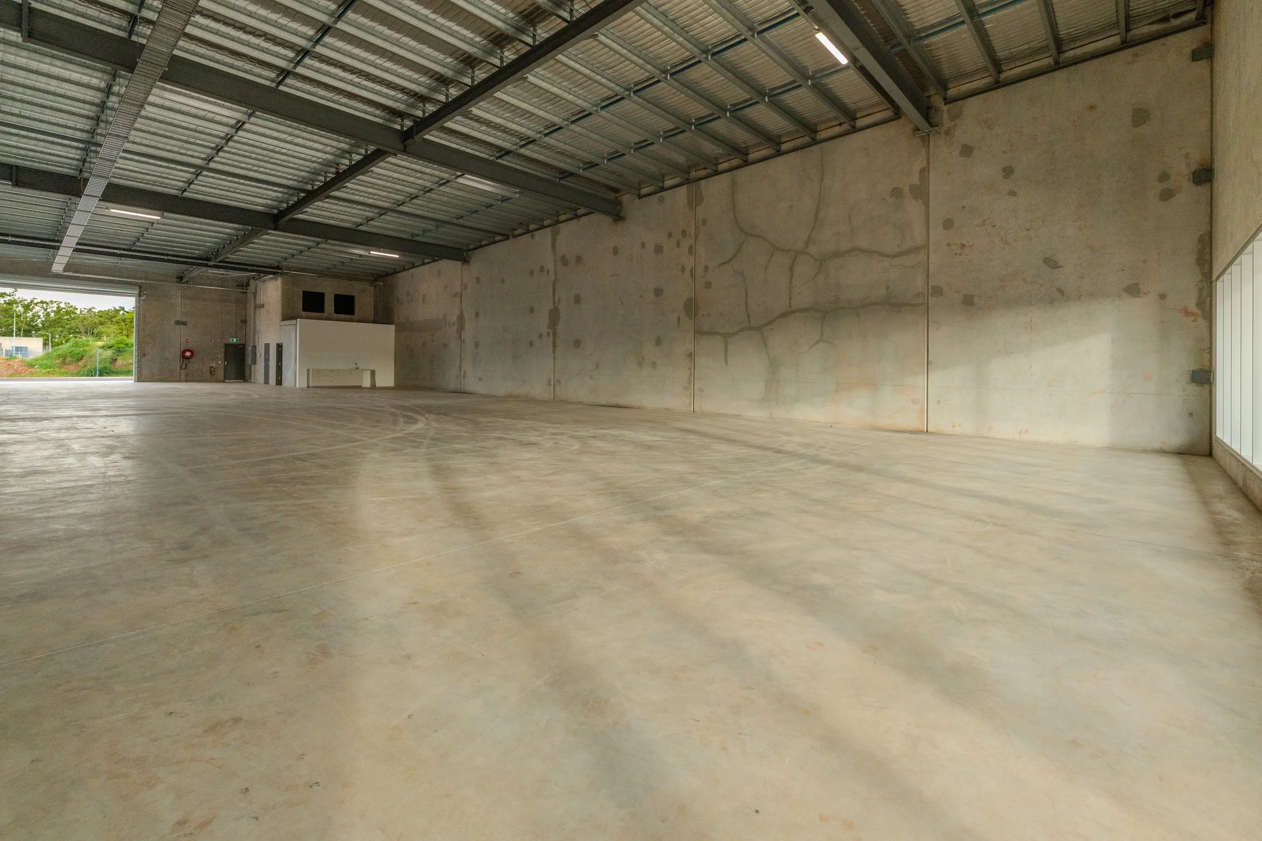 Empty industrial warehouse with concrete floor and metal roof, with some wall cracks, and a large open door showing greenery outside.