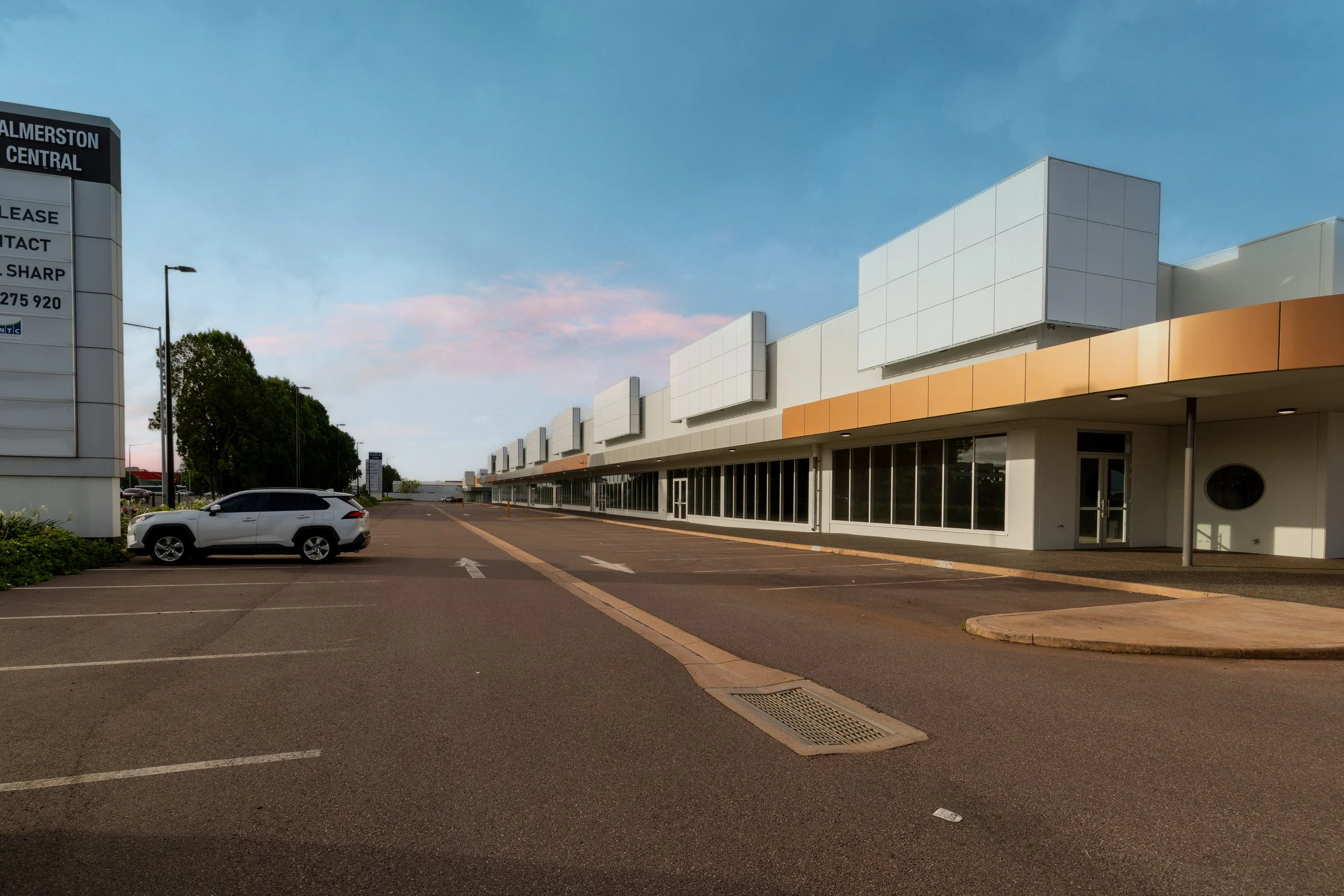 Empty parking lot in front of a modern shopping mall with large glass windows and a white facade under a clear blue sky.