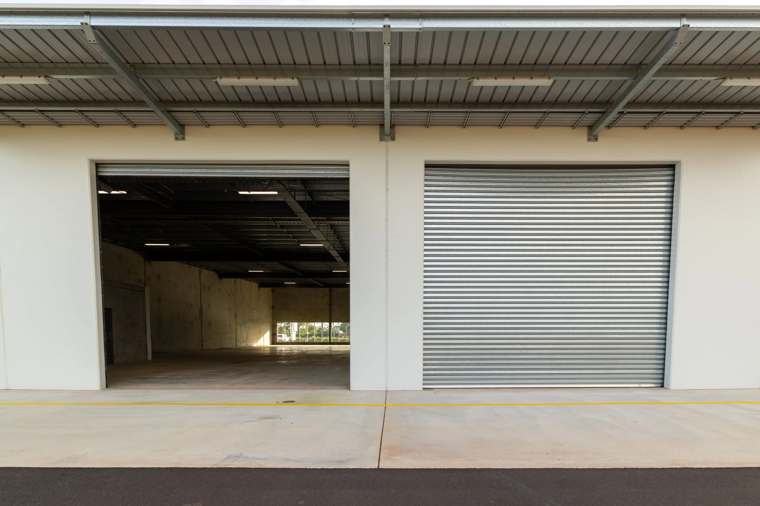 A commercial building with two garage doors, one open showing an empty interior, and one closed with a metal rolling shutter, outside paved area.