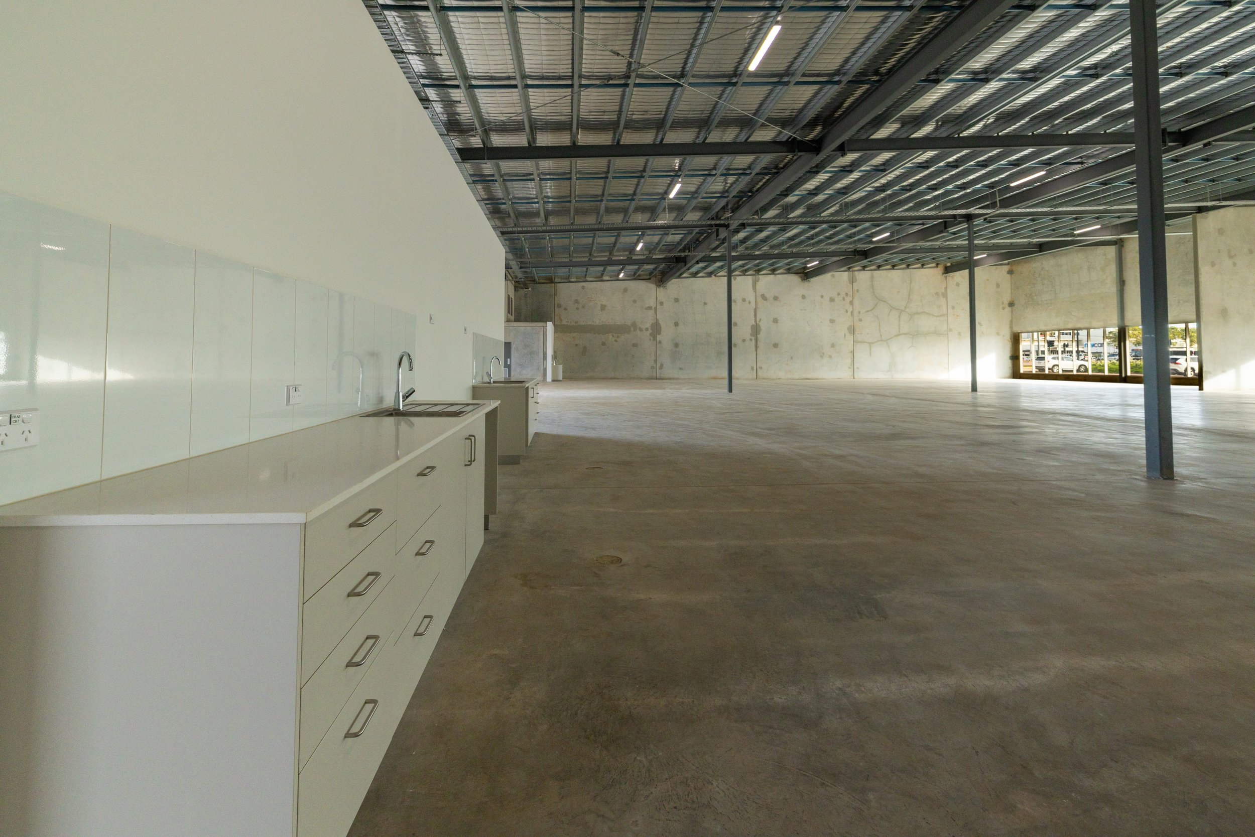 An empty commercial space with unfinished walls, a concrete floor, a kitchen area with white cabinets and sinks, and a high ceiling with exposed metal beams.