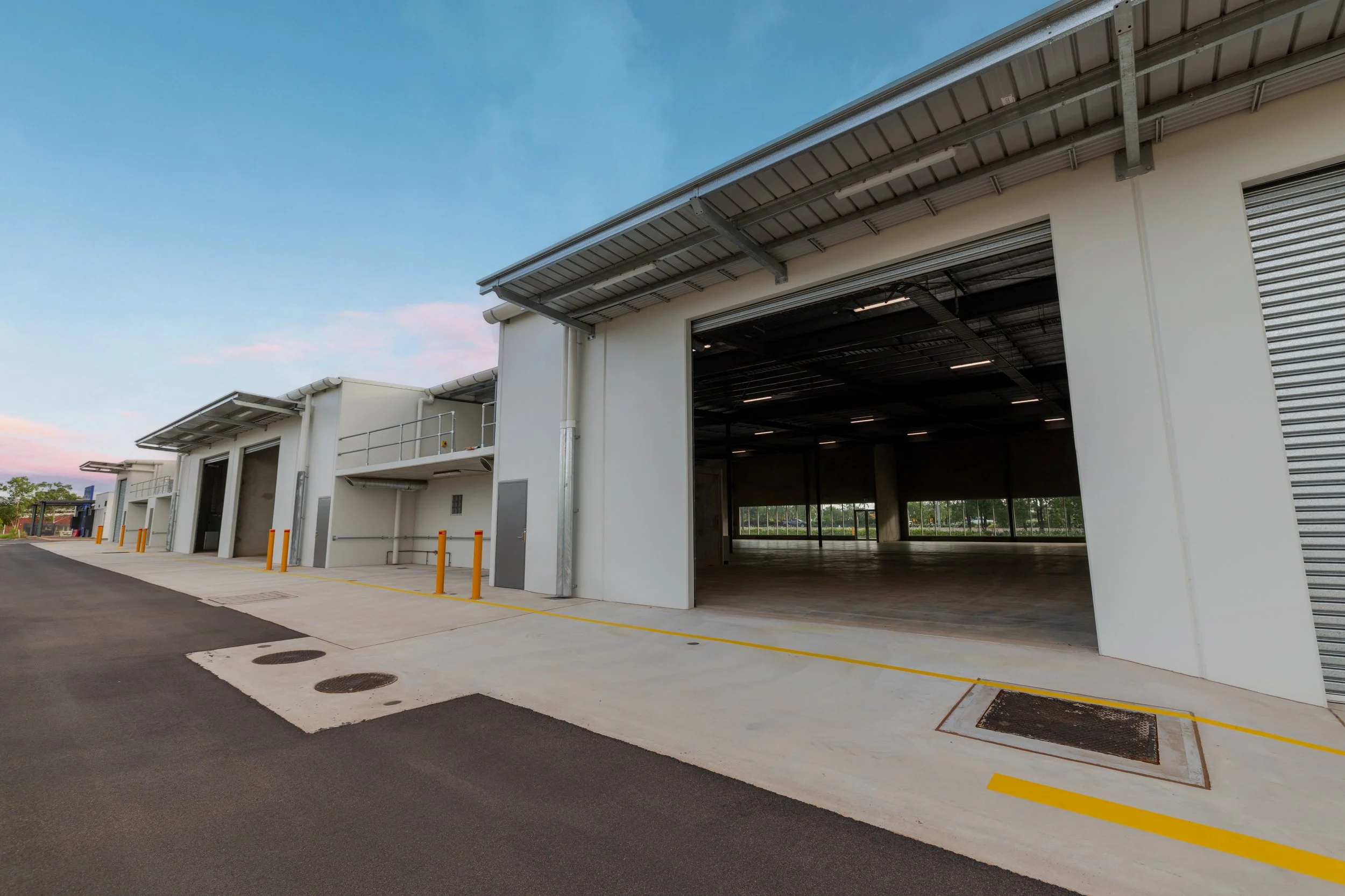 Modern warehouse with multiple open bays, concrete pavement with yellow safety lines, and a clear blue sky.