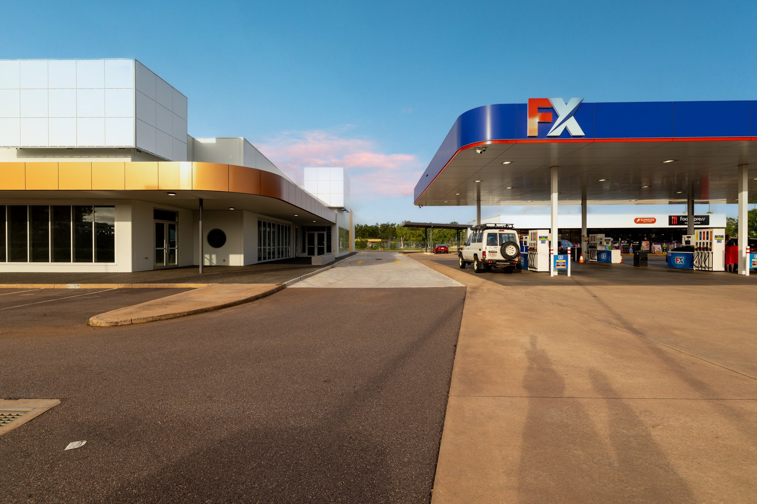 A gas station with a blue and red canopy, fueling pumps, and a small white SUV parked nearby. Modern building with large windows on the left side.