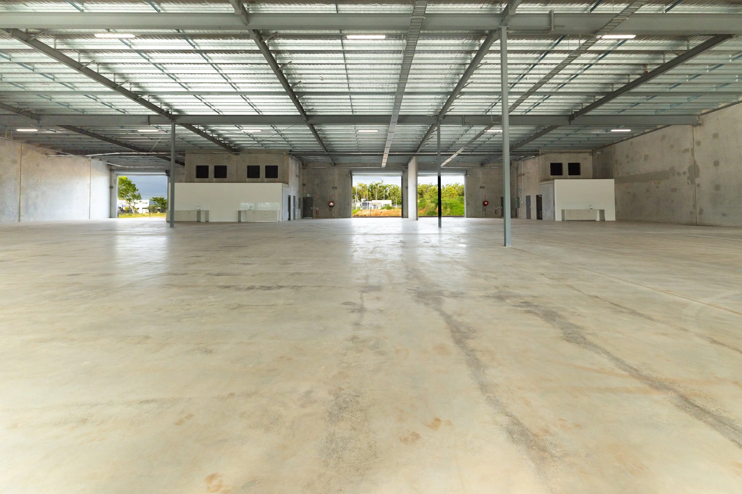 Empty large warehouse with concrete floor, metal ceiling beams, and large open garage doors showing outdoor greenery.