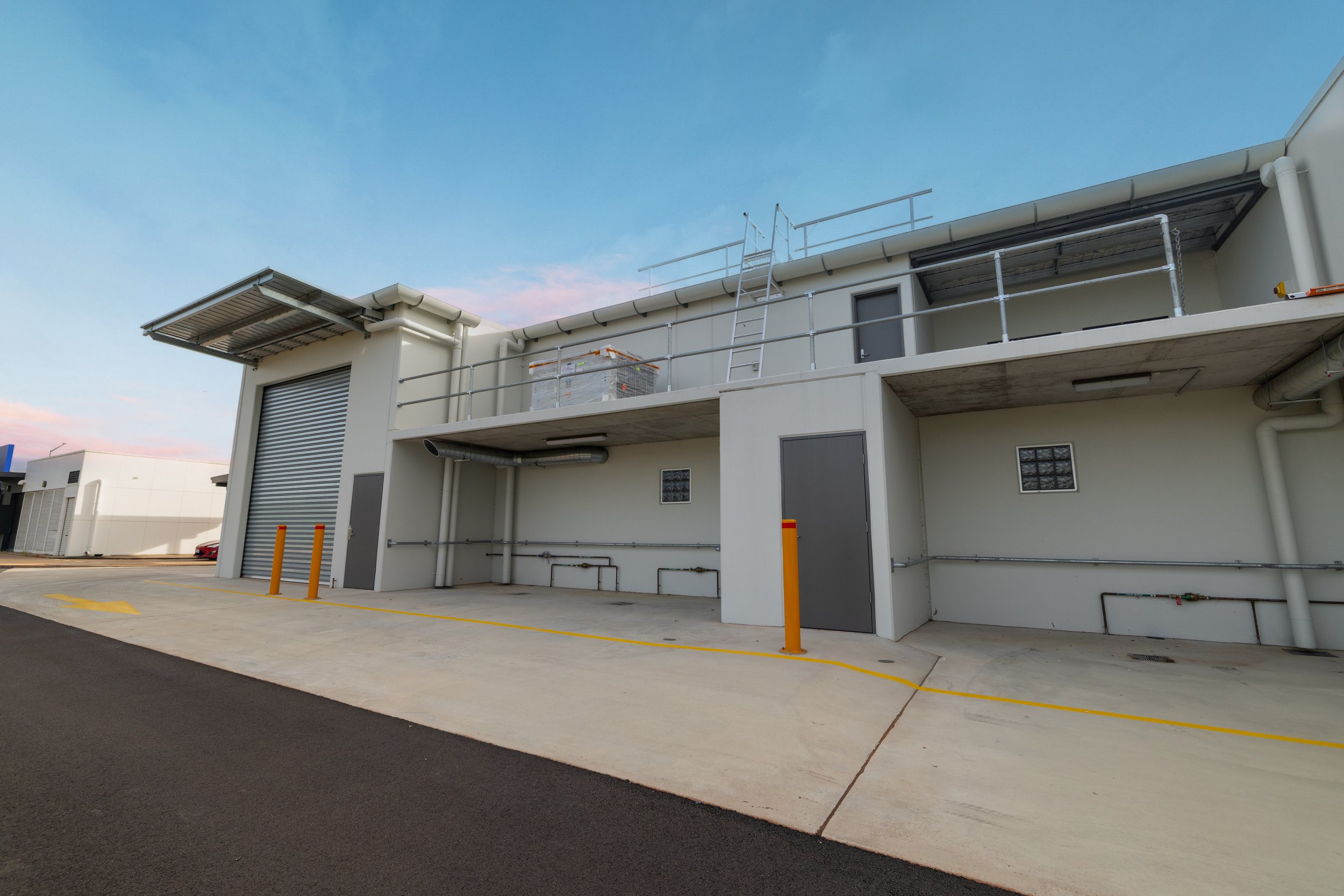 Modern commercial building with a gray roller shutter door, an adjacent gray pedestrian door, and a second-floor balcony with metal railing and ladder, under a blue sky.