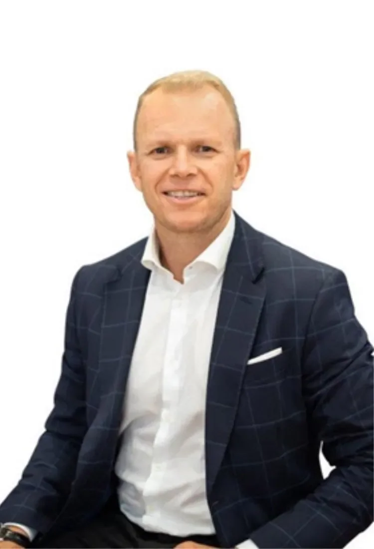A man with short blonde hair and light skin, wearing a dark checkered blazer and a white shirt, is smiling in front of a plain white background. Peter Haverhoek real estate agent based in Adelaide.