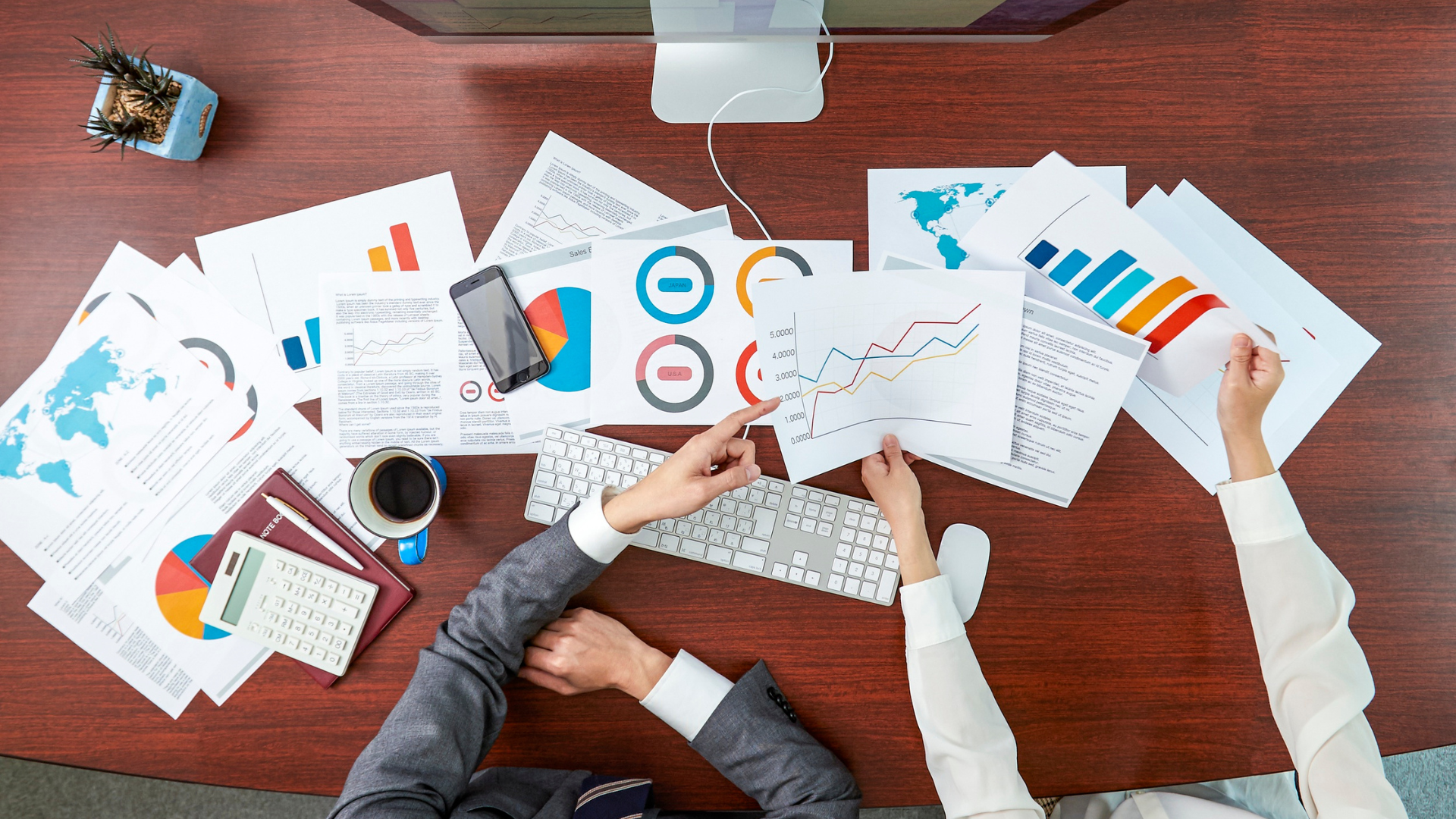A busy office desk with graphs, charts, a calculator, a smartphone, a keyboard, a cup of coffee, and two people analyzing data.