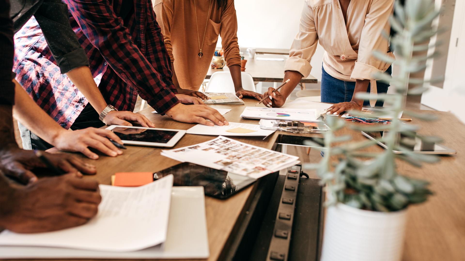 Group of people gathered around a wooden table reviewing documents, tablets, and charts in a meeting room.