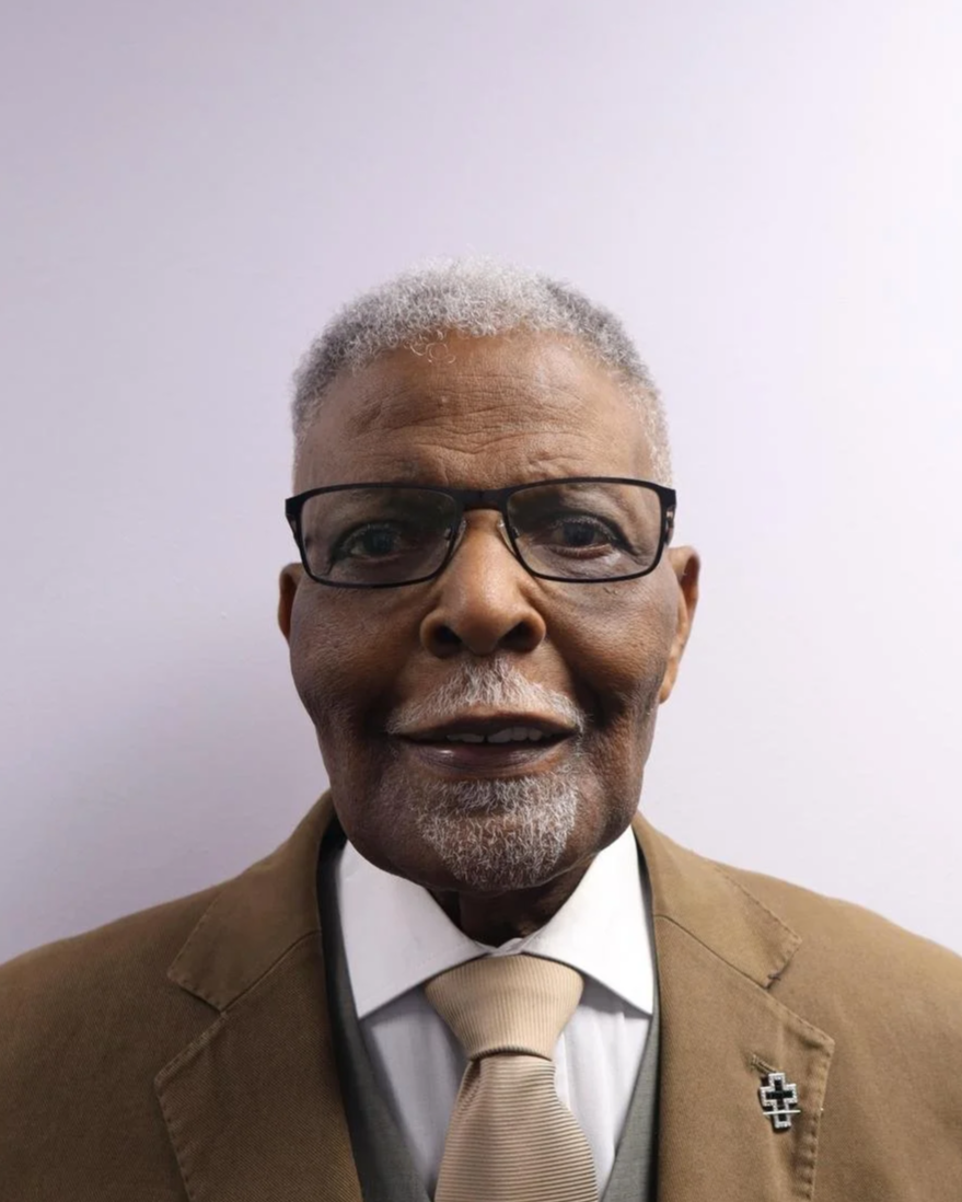 Portrait of an older African American man with gray hair and beard, wearing glasses, a brown suit jacket, a white shirt, and a beige tie, smiling in front of a plain light-colored background.