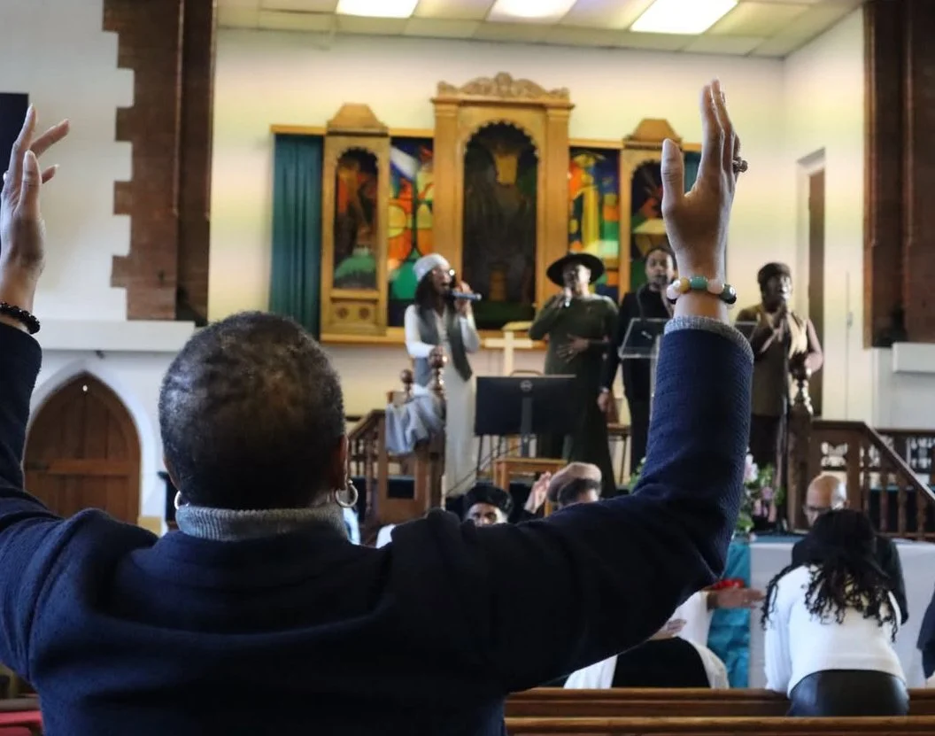 People in a church raising their hands during a service or choir performance with a group of singers on stage.