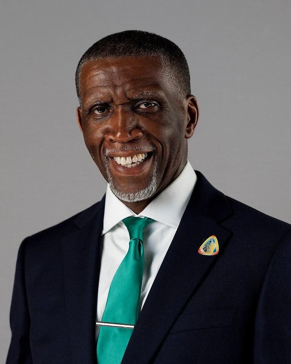 A smiling black man in a dark suit, white shirt, and teal tie, with a pin on his lapel, standing against a gray background.