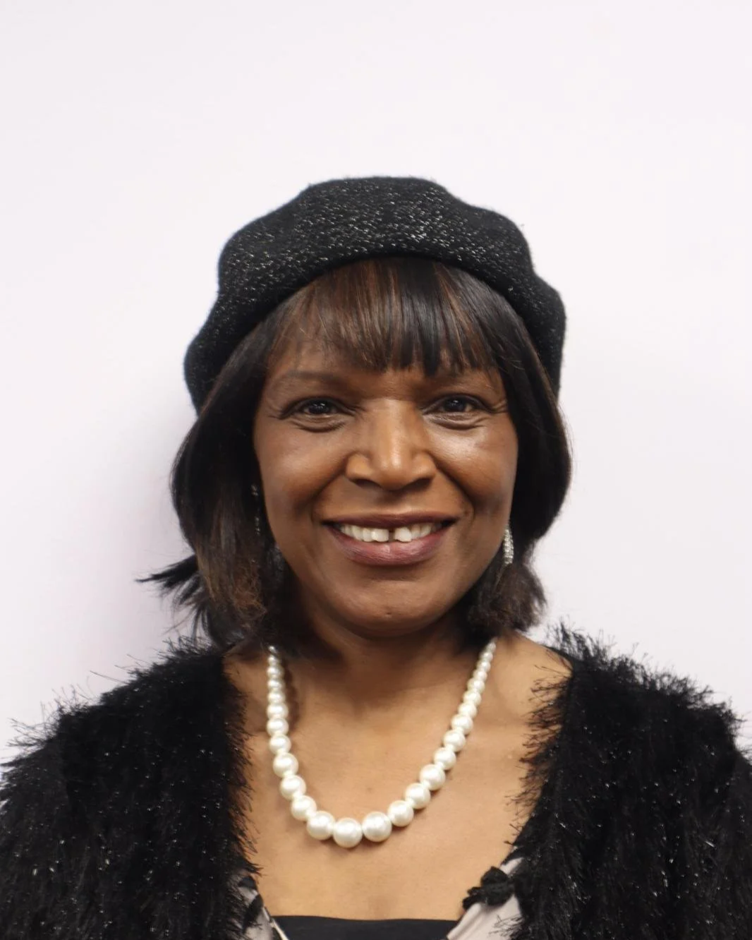 A woman smiling, wearing a black beret, pearl necklace, black fuzzy cardigan, and earrings, in front of a white background.
