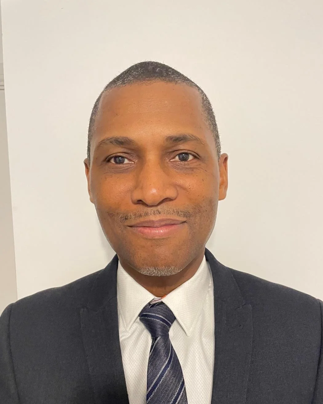 A man in a black suit, white shirt, and striped tie smiling in front of a plain white wall.