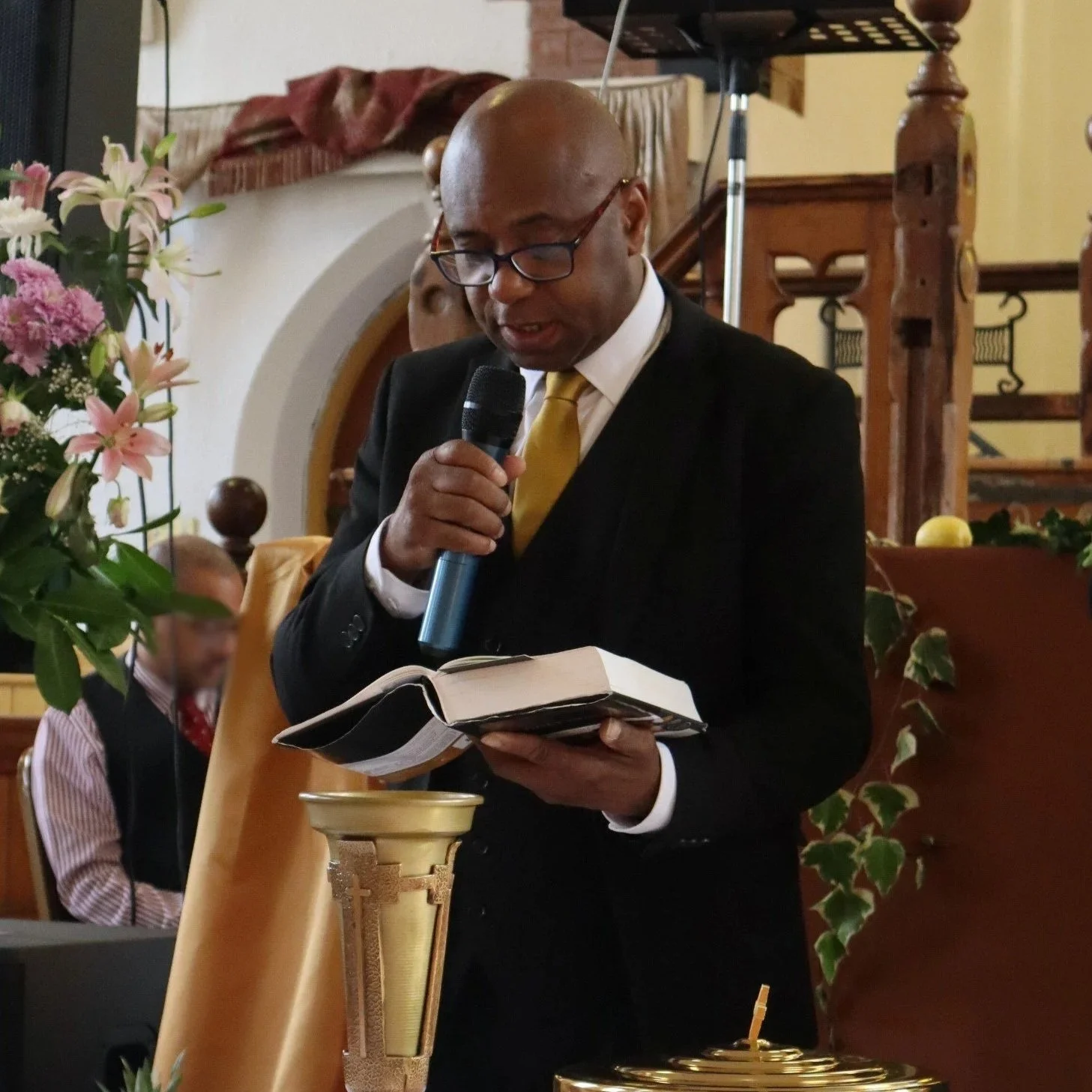 A man dressed in formal attire, wearing glasses and a yellow tie, is speaking into a microphone while holding an open book. He is standing behind a gold-colored stand or lectern in a decorated indoor setting with flowers and wood paneling.