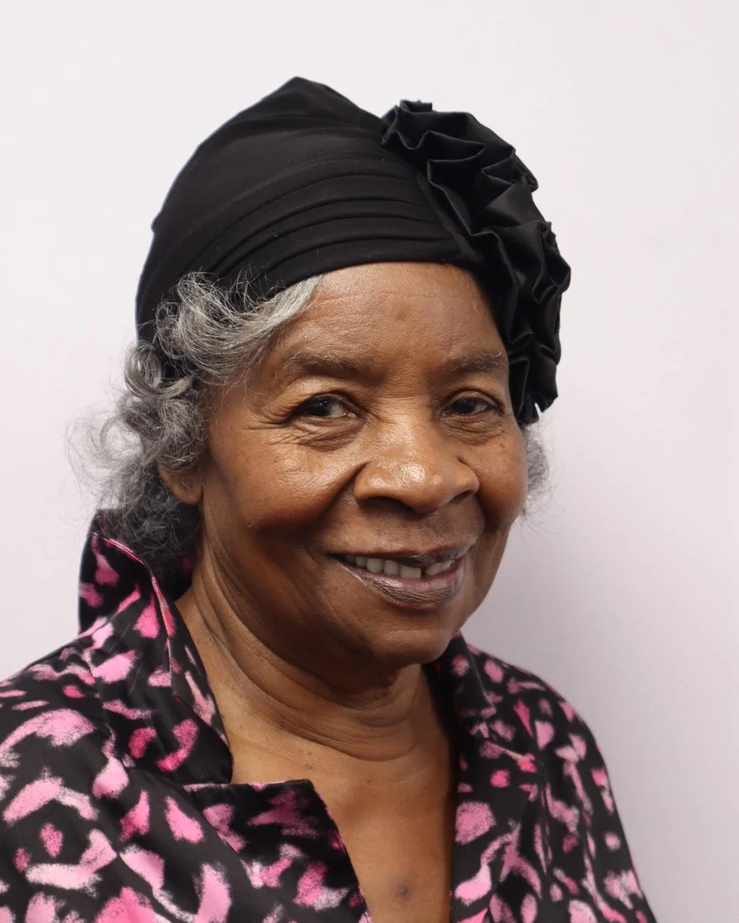 A smiling elderly woman with gray hair wearing a black headscarf and a pink and black patterned blouse.