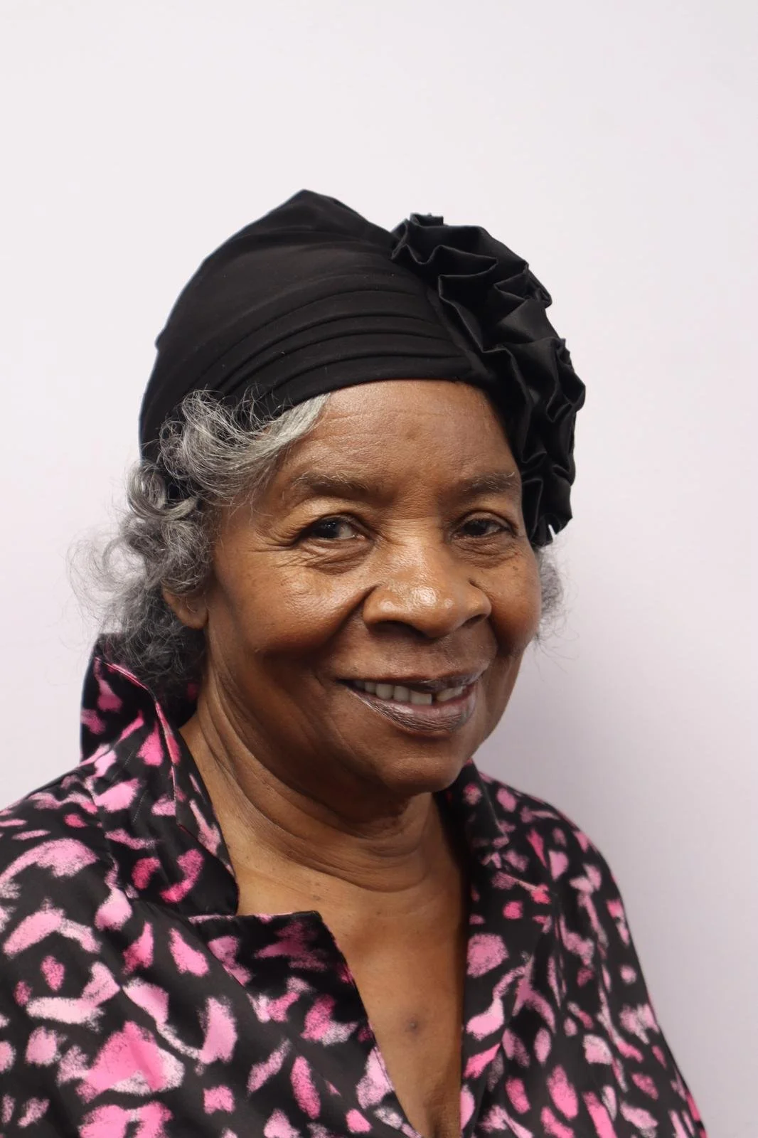 A smiling elderly woman with gray curly hair wearing a black headwrap and a black and pink animal print blouse against a plain white background.