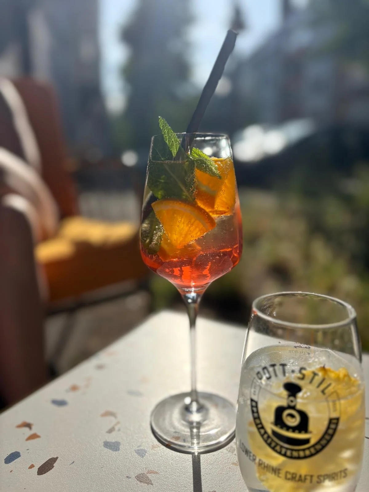 Ein bunter Cocktail mit Minzblättern und Orangenscheiben in einem hohen Glas, neben einem Glas mit alkoholischer Spirituose auf einem weißen Tisch im Sonnenlicht.