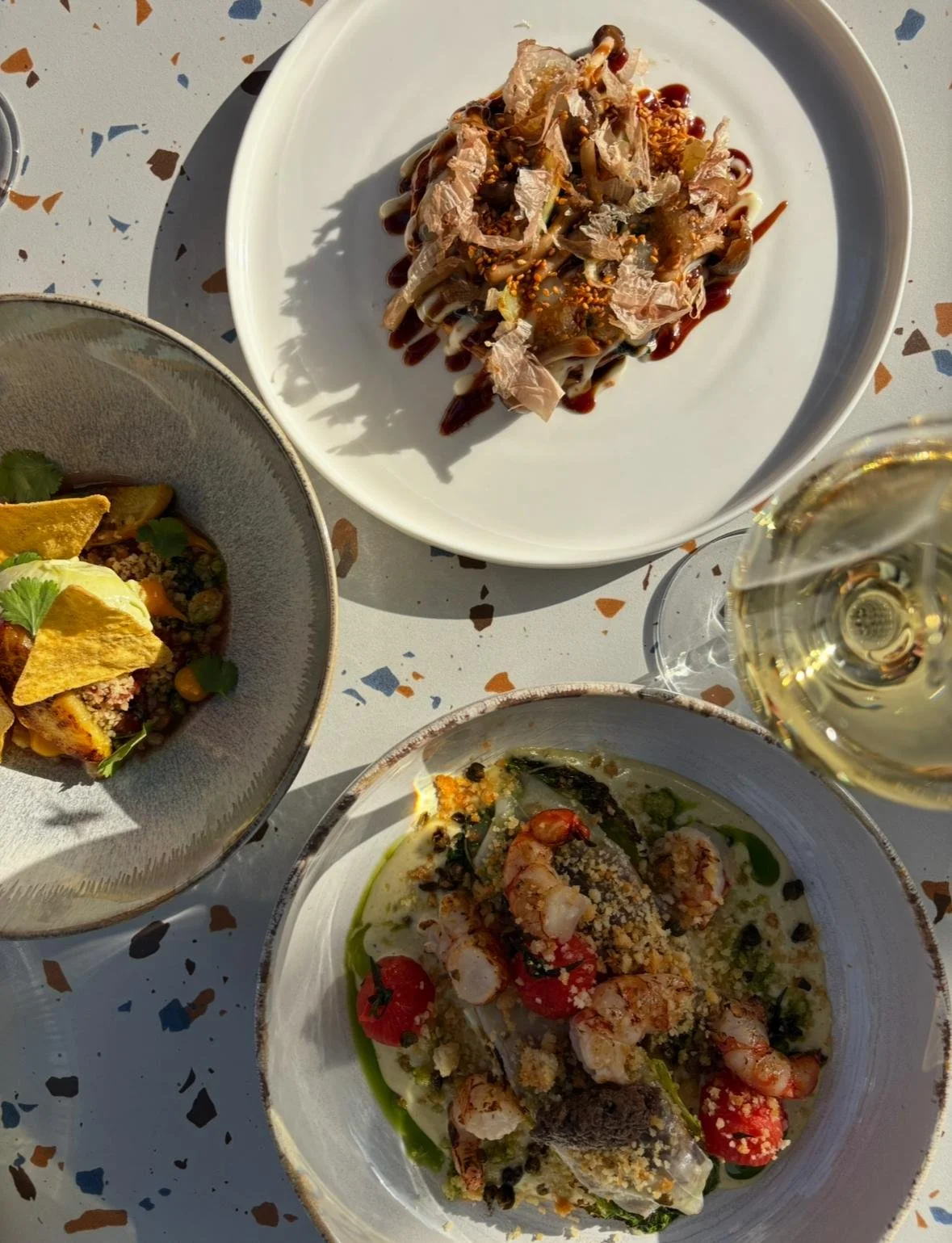 Verschiedene Teller mit Gourmet-Gerichten und ein Glas Weißwein auf einem bunten Tisch.