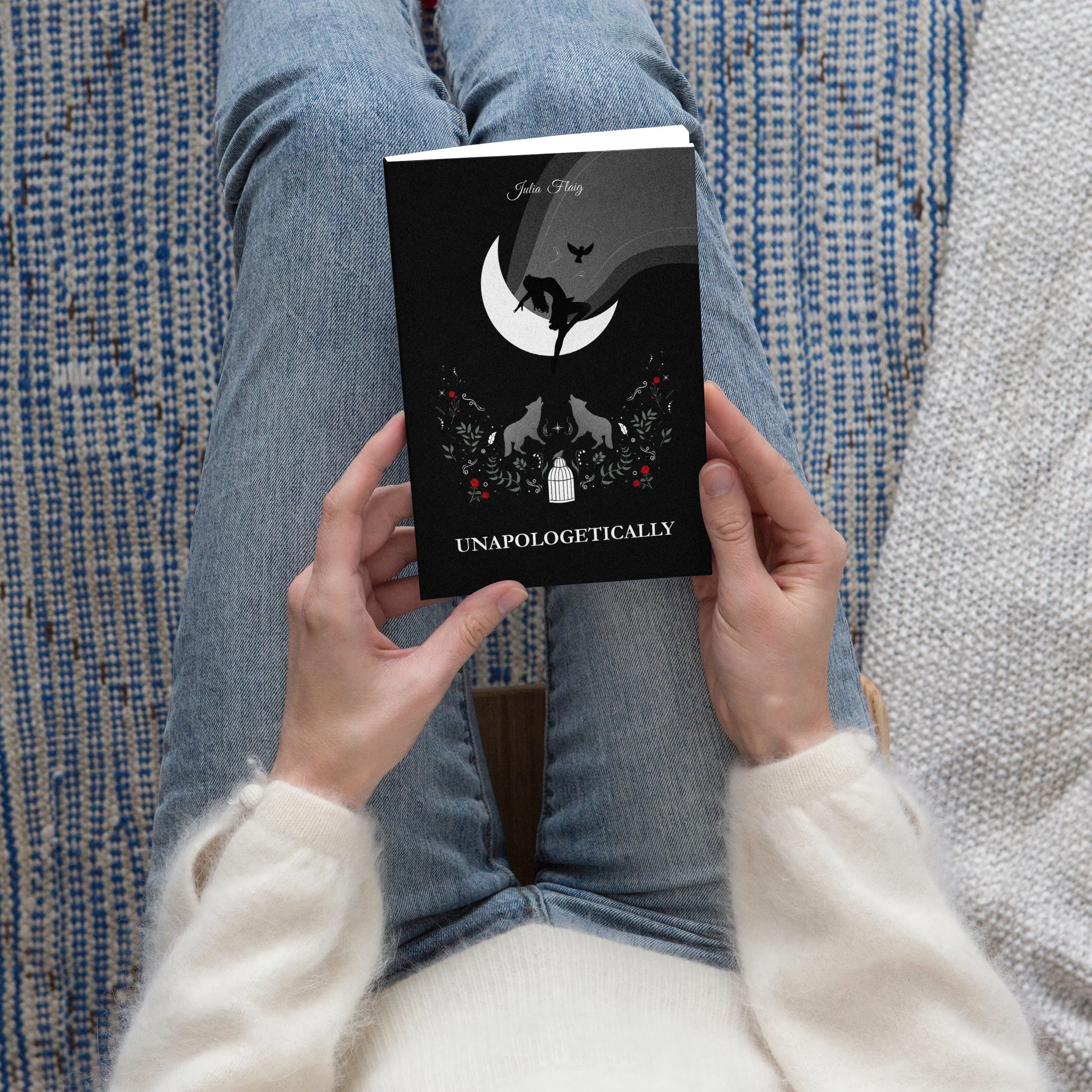 Person holding a black book titled 'Unapologetically' while sitting on a patterned blue and white sofa, wearing a white sweater and jeans.