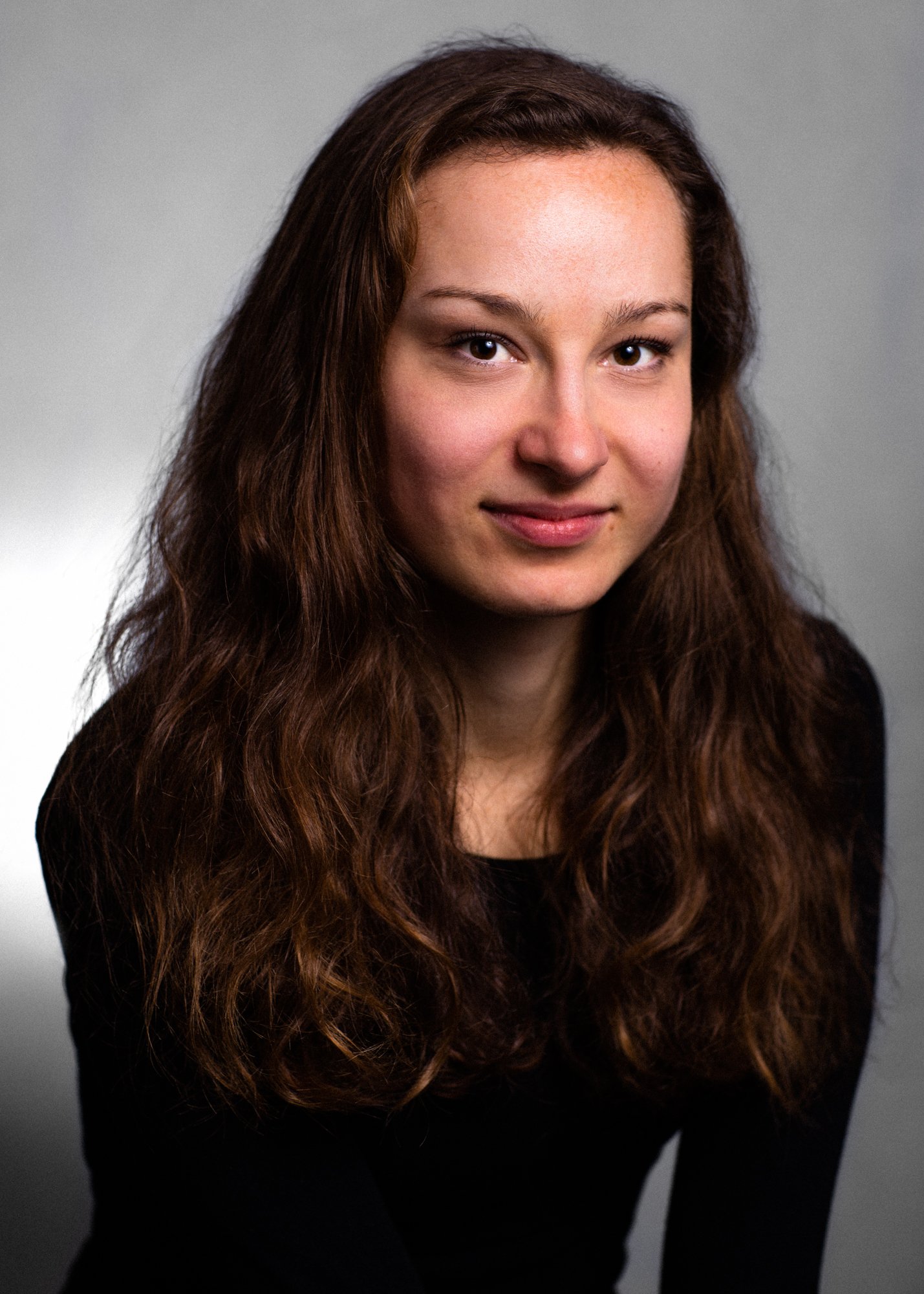 A portrait of a woman with long, wavy brown hair, wearing a black top, looking at the camera with a slight smile against a gray background.