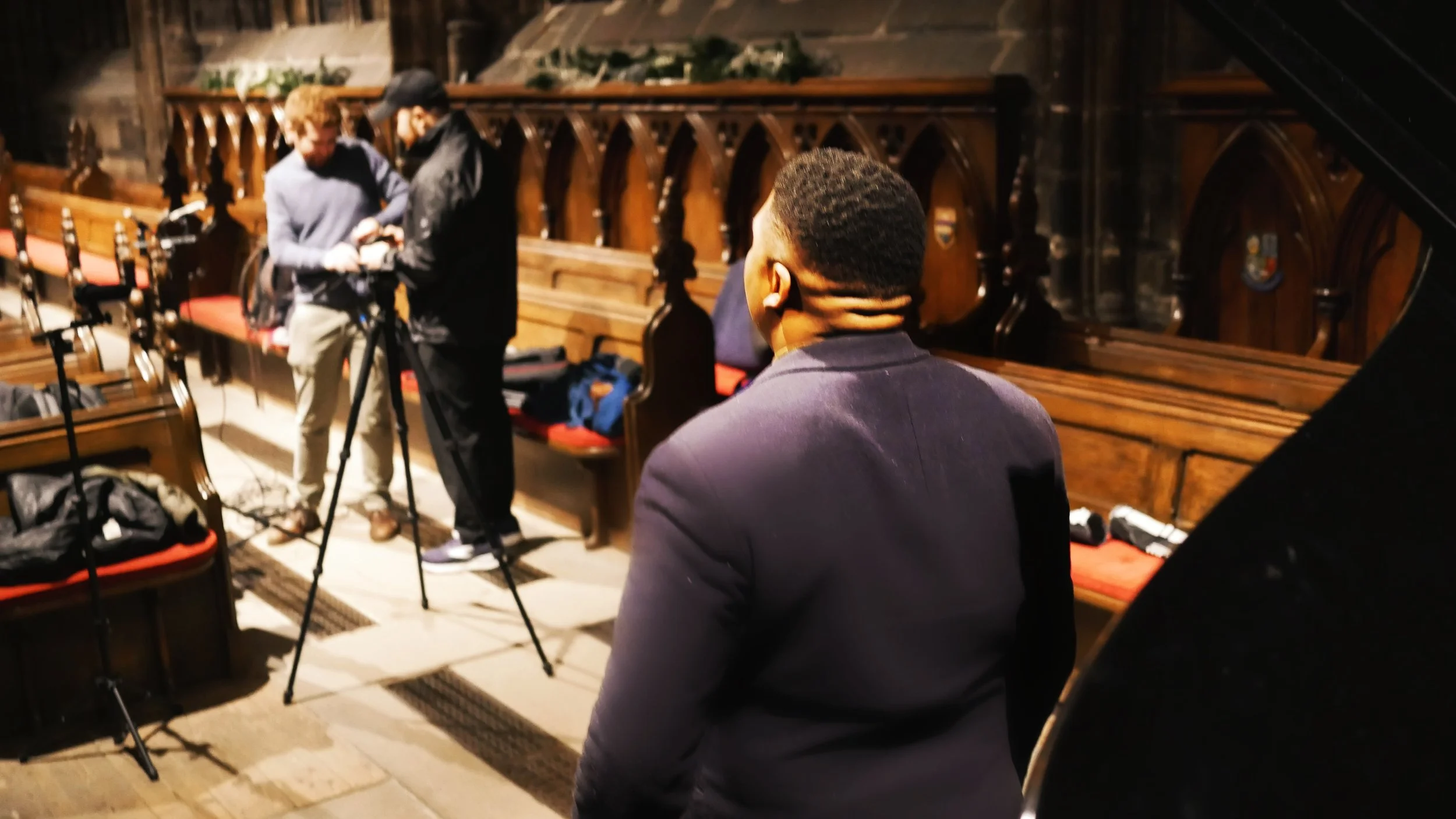 Man in a dark suit observing a camera crew in a church with wooden pews and gothic architecture.